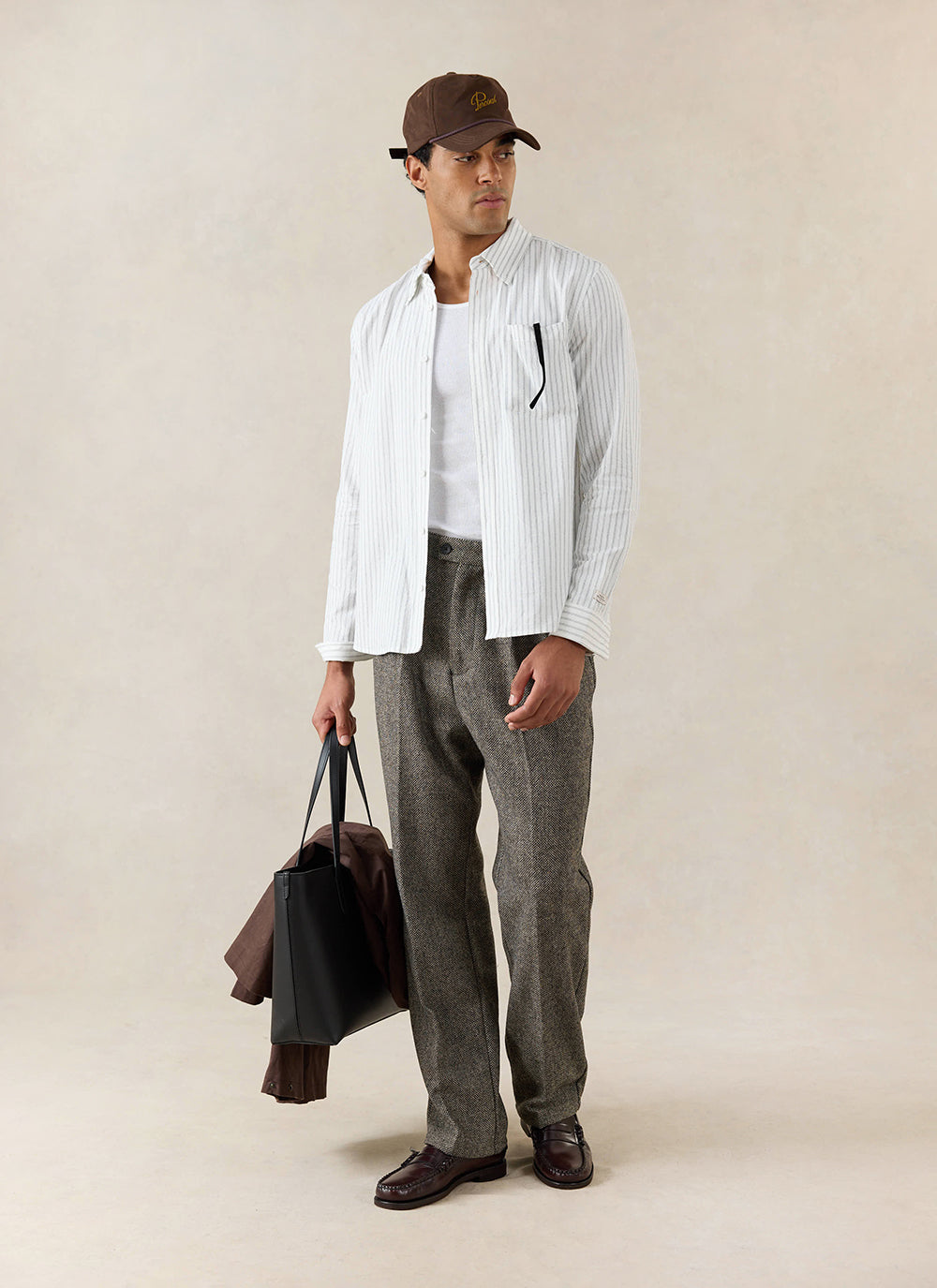 A model wears the white Percival Fabian Striped Shirt in cotton, styled with grey trousers and a brown baseball cap.