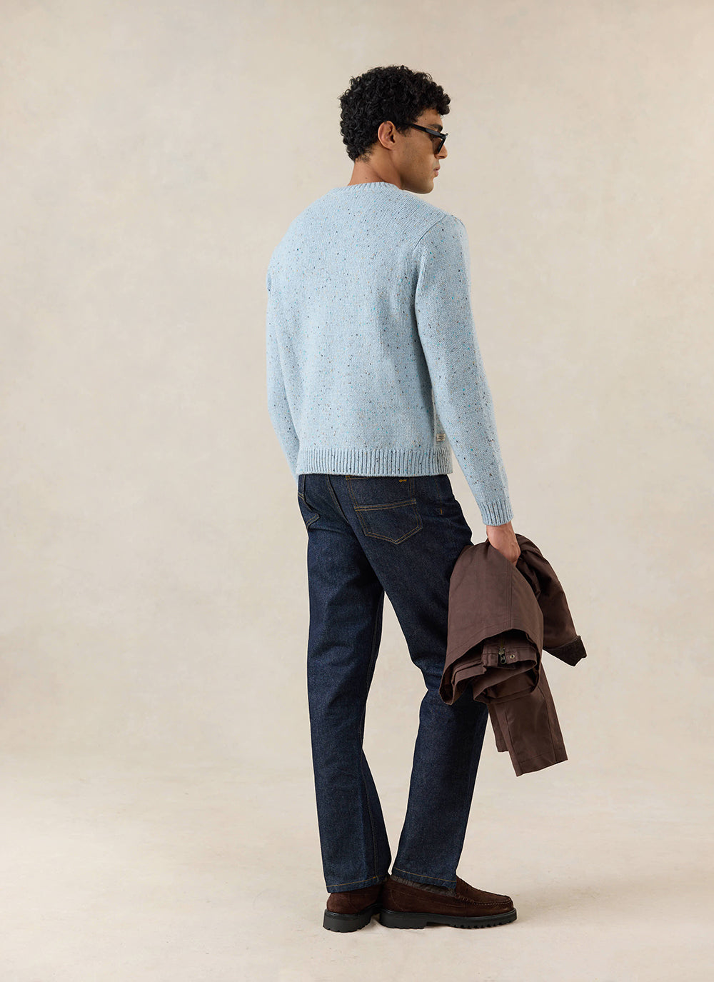 A model wears the blue Percival Rhodes crew neck jumper, styled from the back with dark denim jeans and a brown jacket.