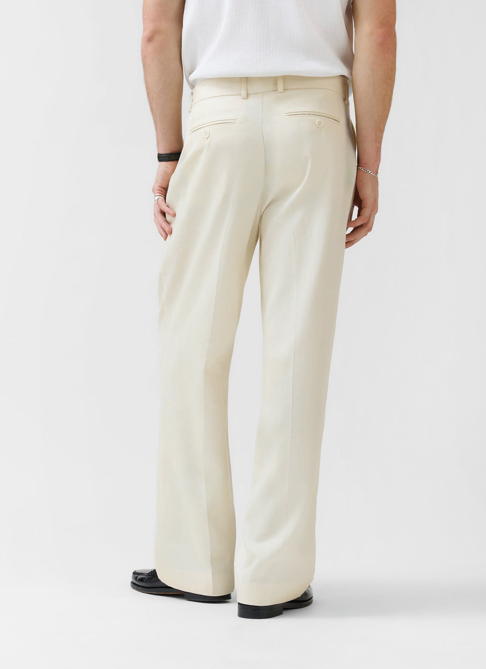 A model, seen from behind, wears the Percival Pleated Tailored Trousers in cream, showing the relaxed fit and drape.