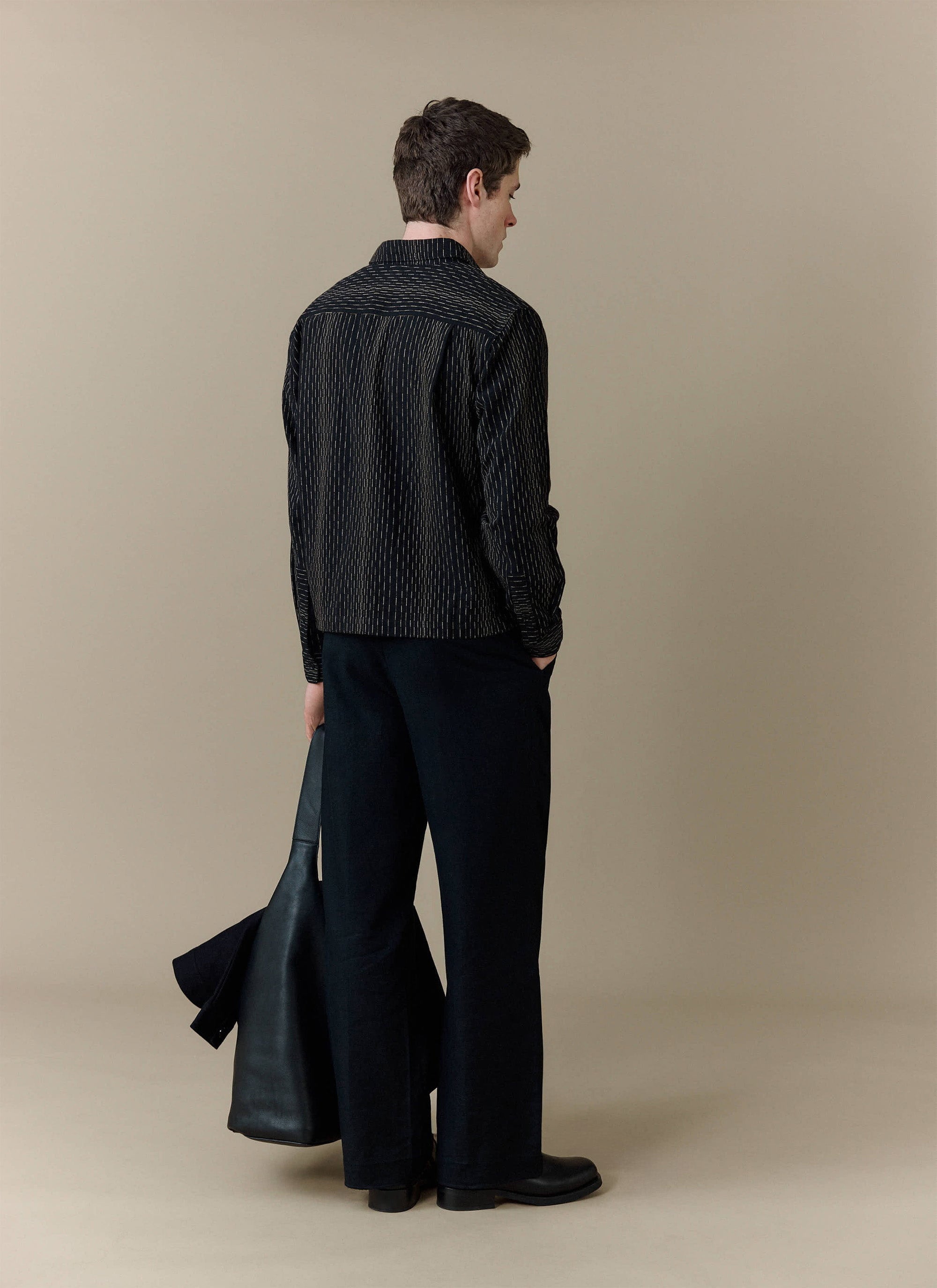 A rear view of a model wearing the black Leland Cuban Boxy Shirt from Percival, styled with black trousers.