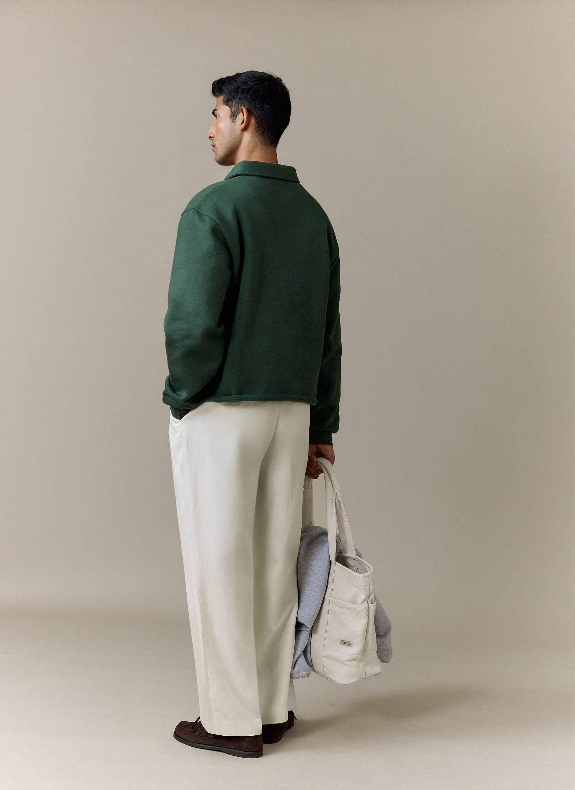 A model seen from behind wearing the Percival Dudley Boxy Sweatshirt in forest green with white trousers and a tote bag.