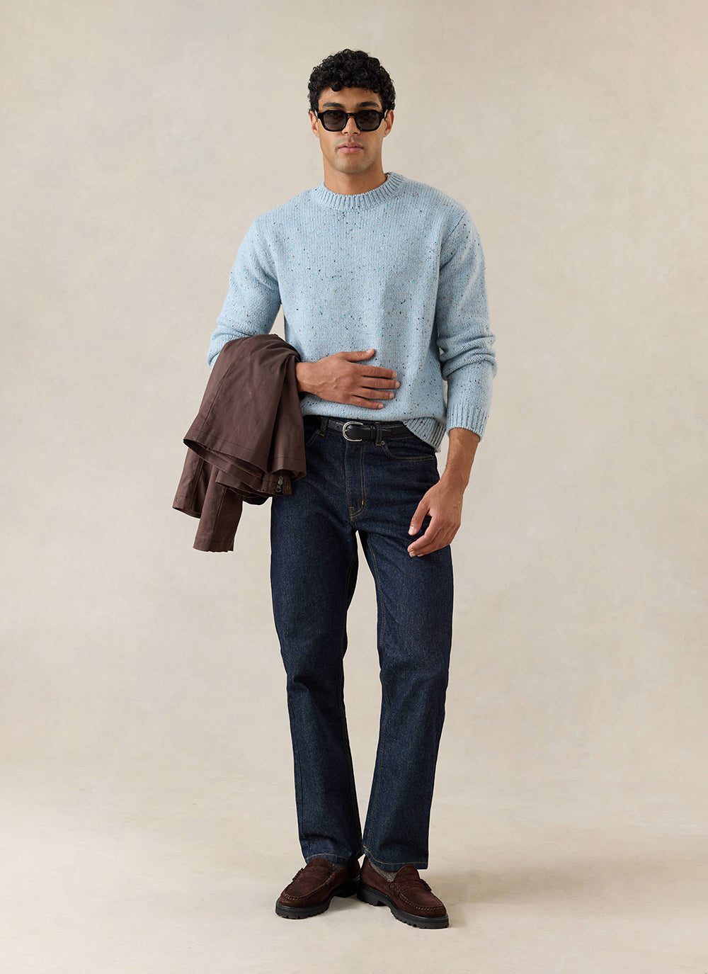A model wears the blue speckled Percival Rhodes crew neck jumper with dark indigo jeans and brown loafers in a studio shot.