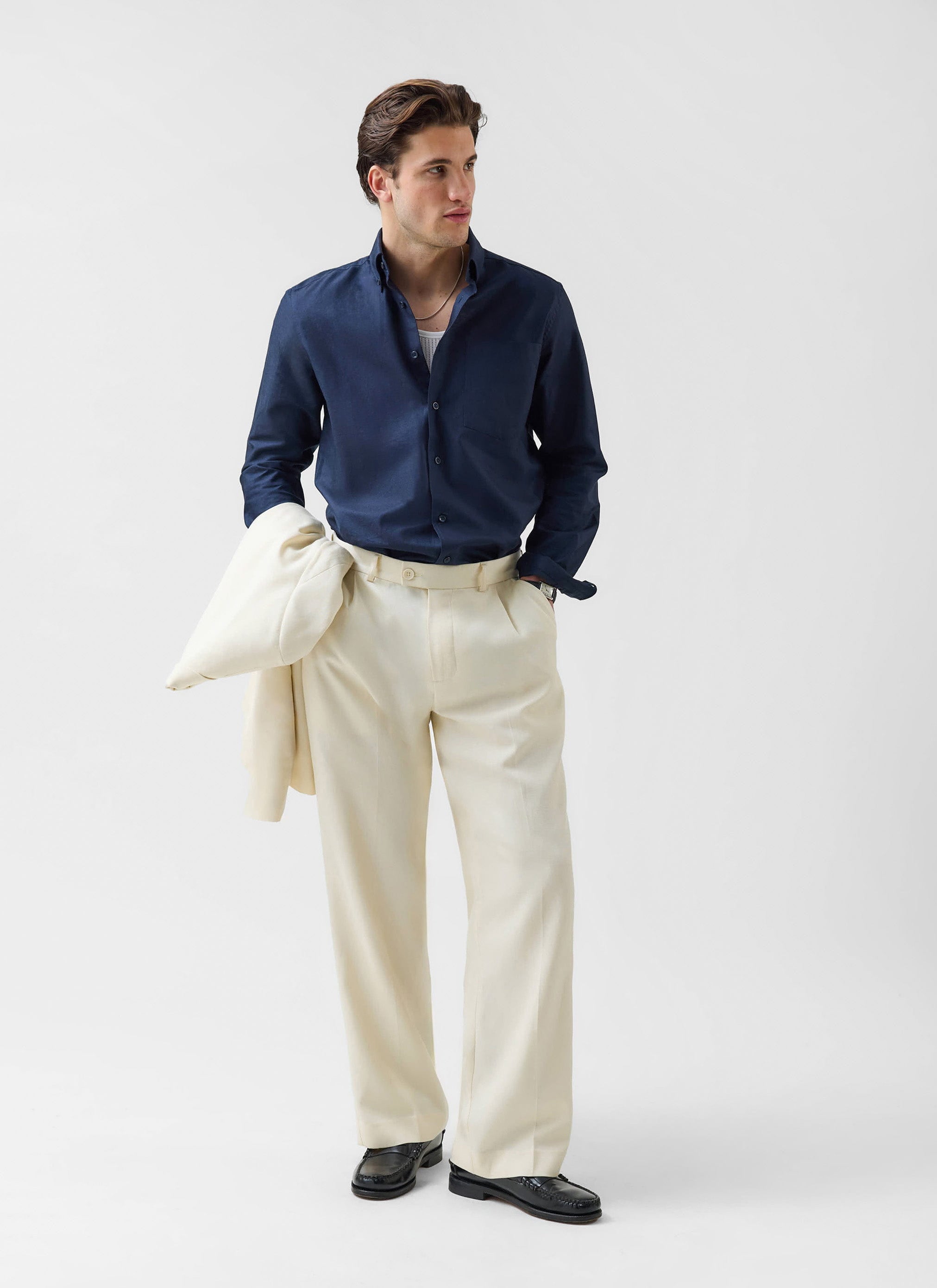 A man models the Percival Pleated Tailored Trousers in cream, styled with a navy shirt and holding the matching blazer.