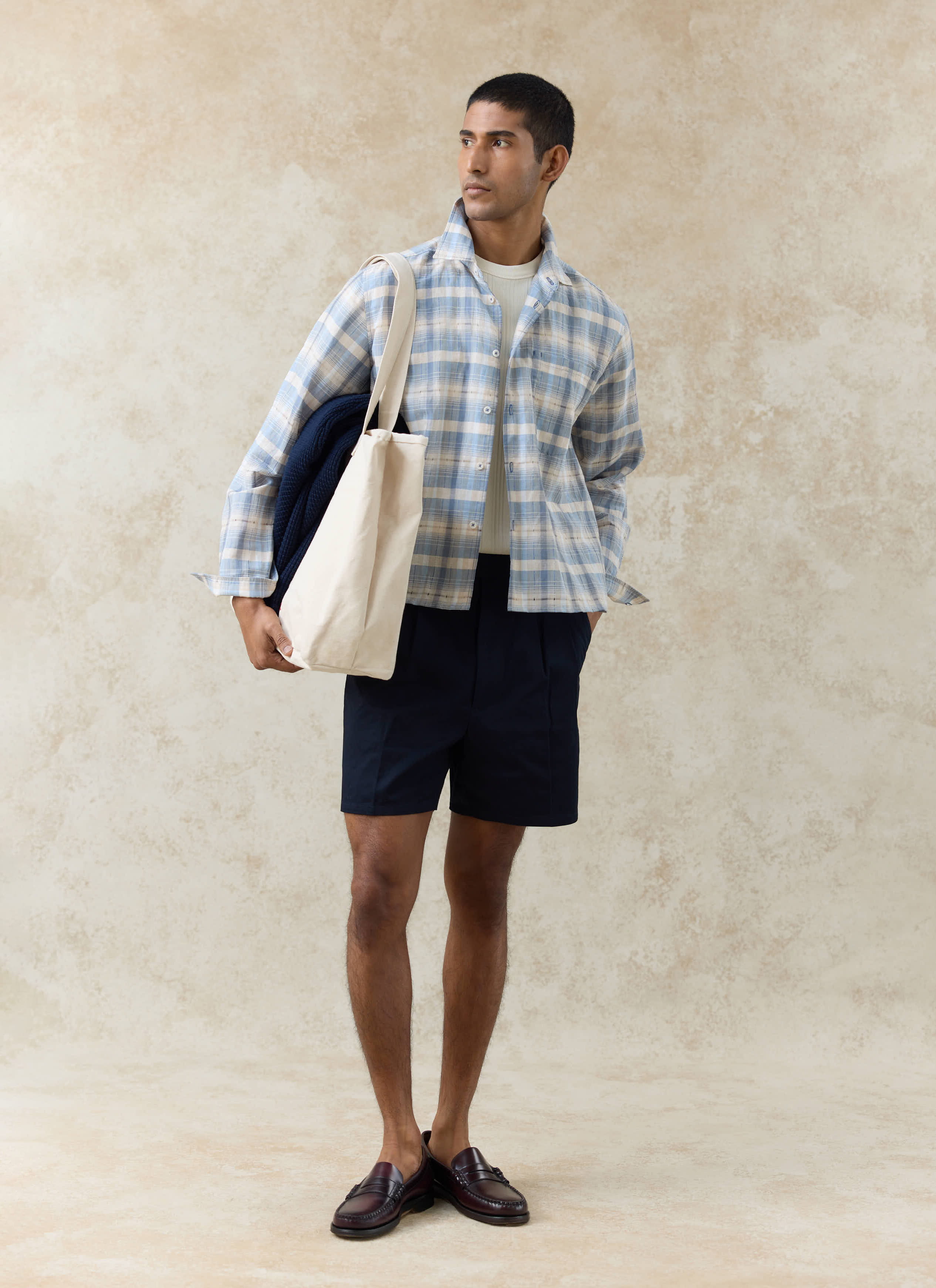 Navy Percival Blake double pleated cotton shorts worn by a model with a blue checked shirt and cream tote bag.