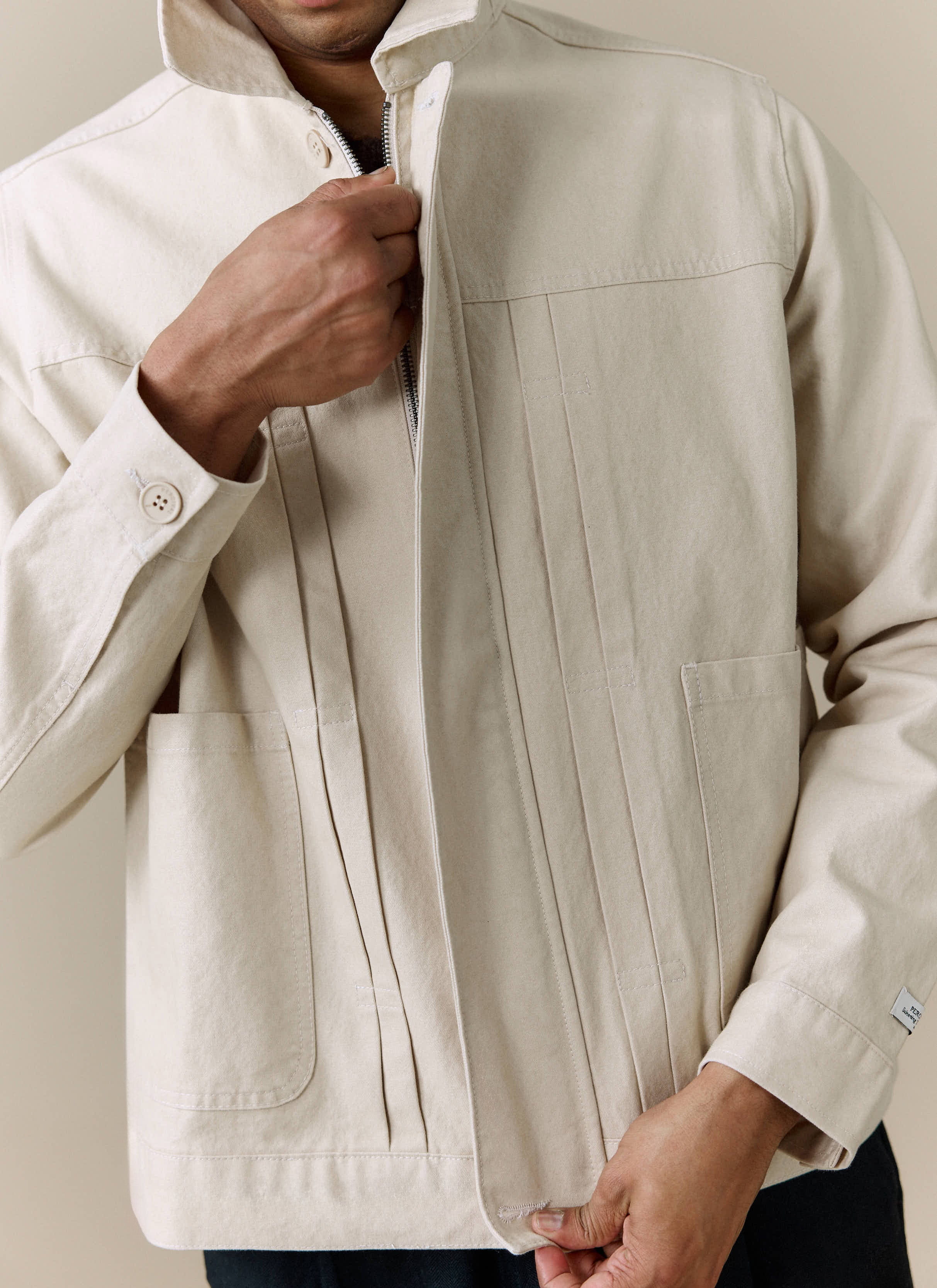 A close-up of a model zipping up the Percival Ernst Jacket in ecru, showing the pleated front detail.
