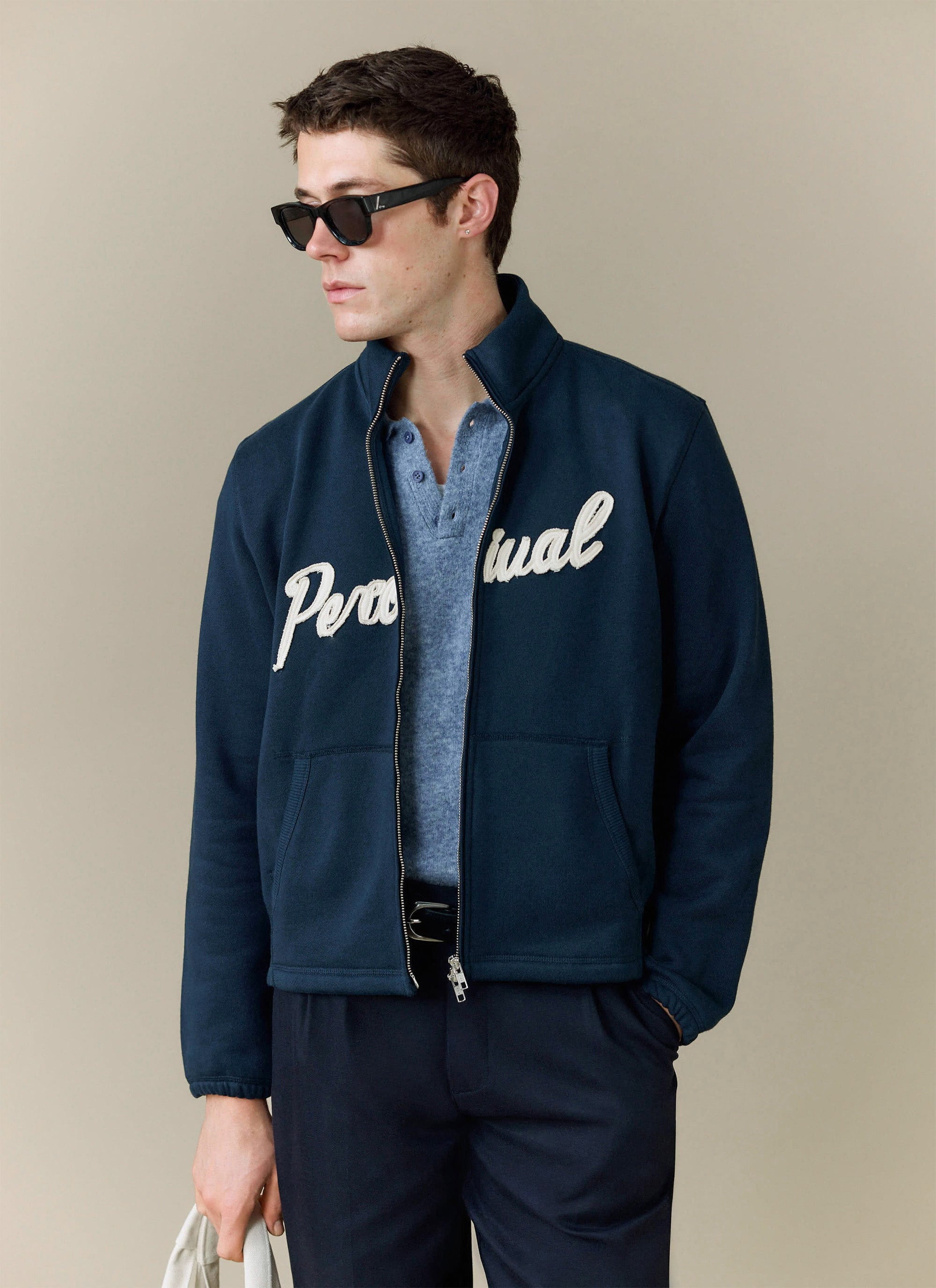A model wears the Percival Collins Knitted Polo in blue, styled under a navy zip-up jacket with Percival branding.