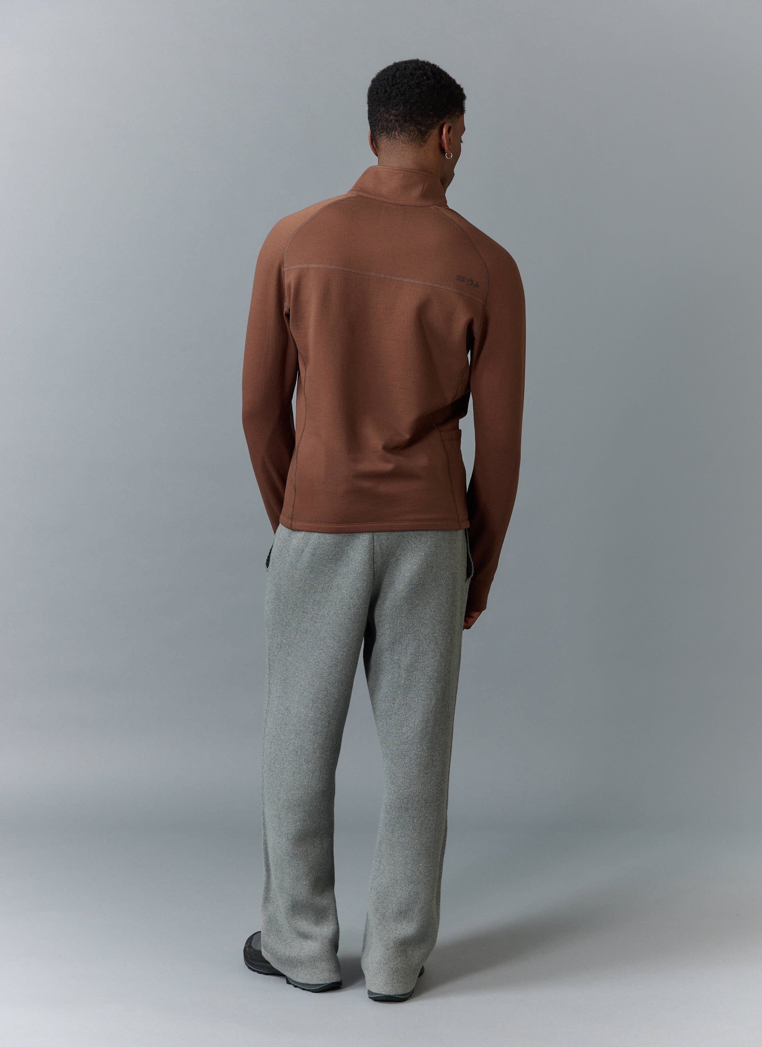 A model from behind wearing the Percival PCVL Base Layer Quarter Zip Jacket in rust, styled with grey trousers.