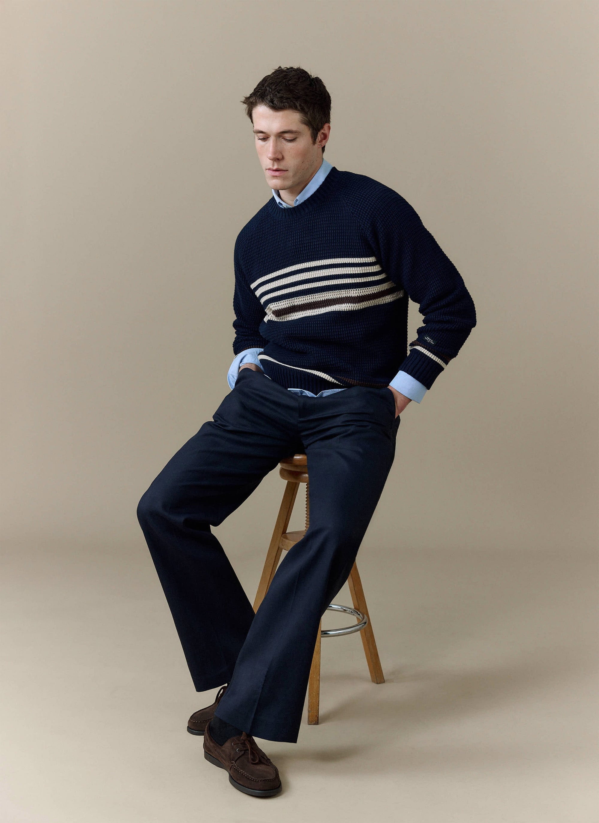 A model sits on a stool wearing the Percival Sergio Crochet Jumper in navy with matching trousers and brown shoes.