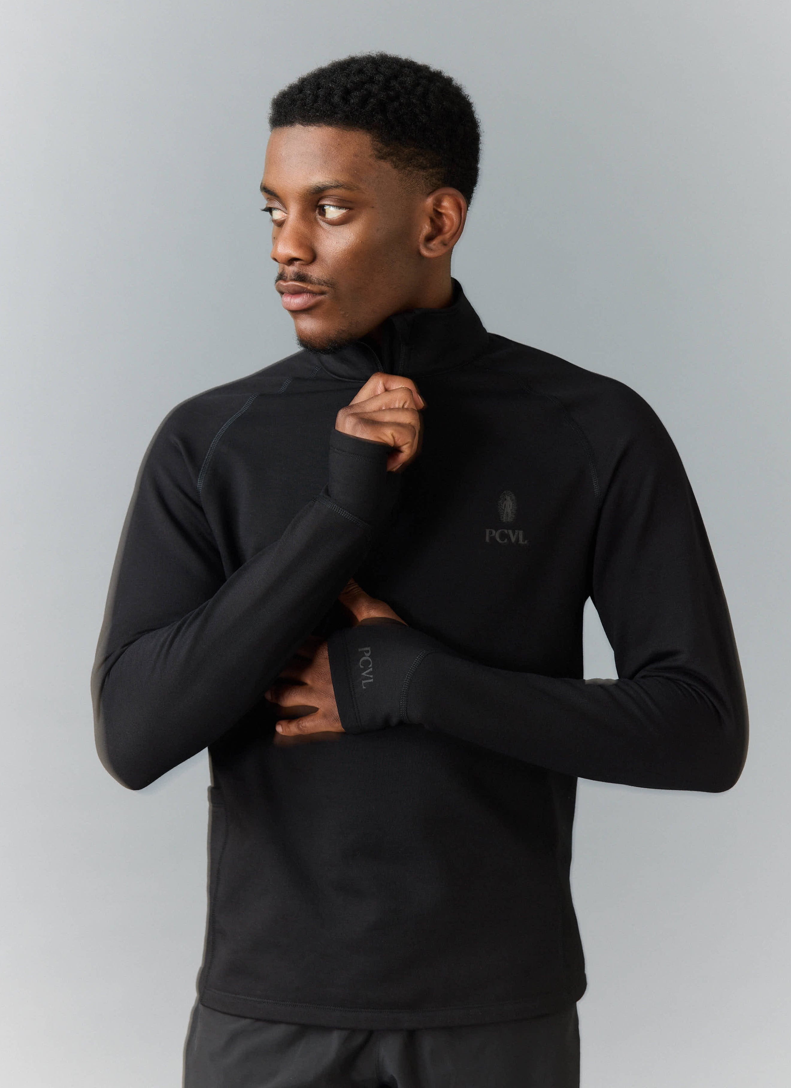 A model wears the black PCVL Base Layer Quarter Zip by Percival, showing the thumbhole cuffs and tonal chest logo.