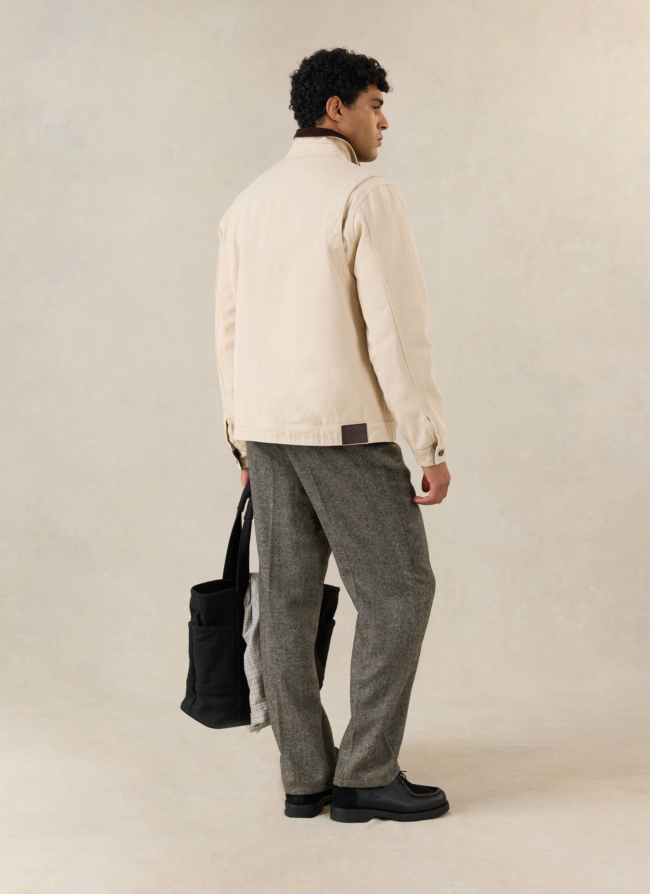 A model wears the cream cotton Percival Fontane Jacket, styled with textured grey trousers and a black tote bag.