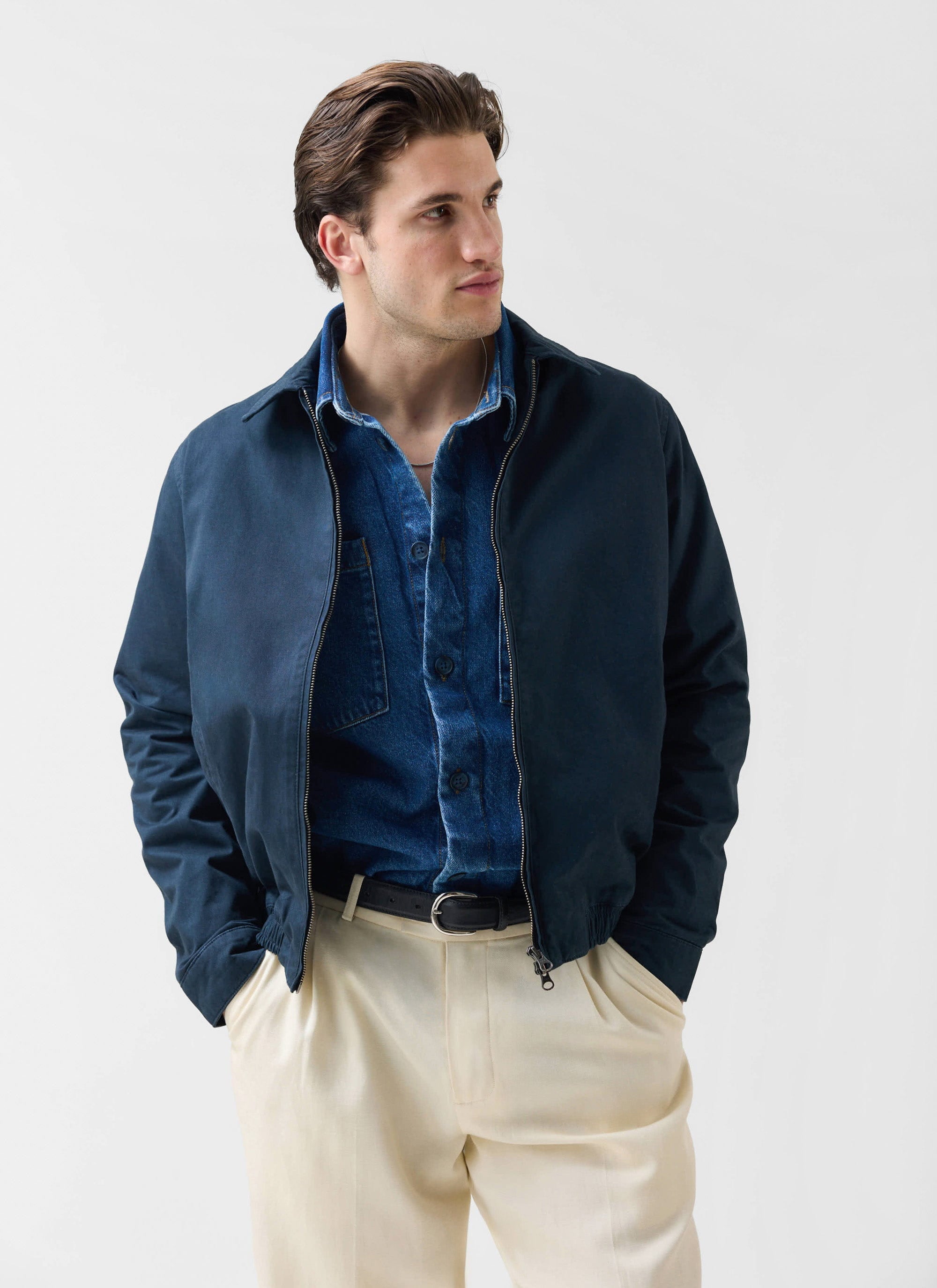 A man models the Percival Leo Harrington jacket in navy, styled with a denim shirt and cream trousers.