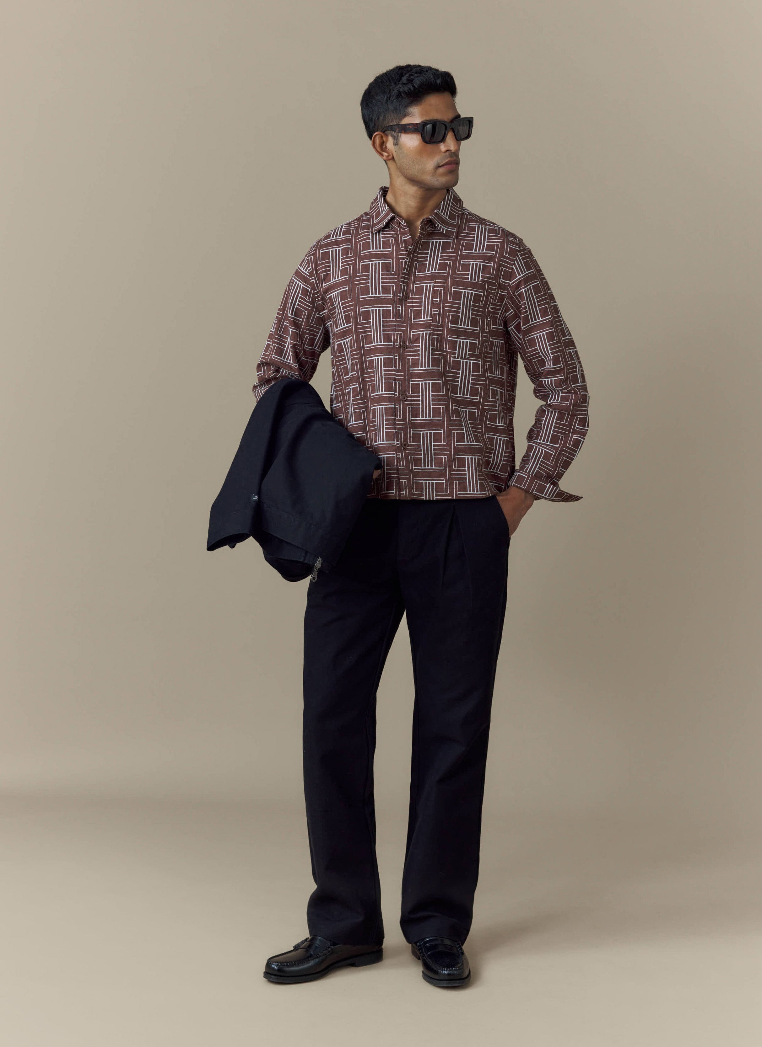A model wears the Percival Sashiko Boxy Shirt in brown with black trousers and sunglasses, holding a dark jacket.