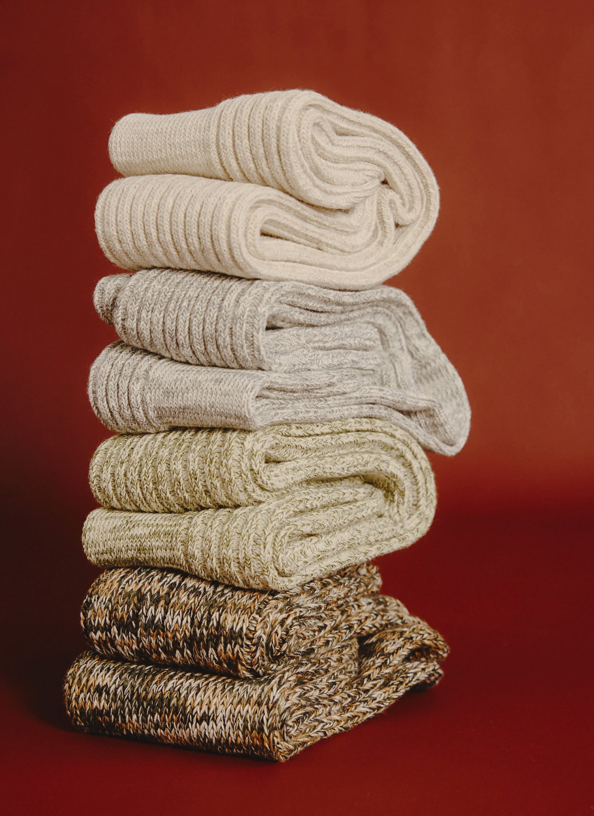 A stack of four pairs of ribbed, marl cotton socks from Percival in neutral colours, set against a rich red background.