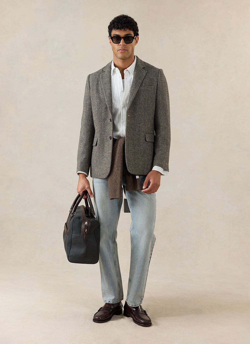 A model wears the Percival Wilde Tailored Blazer in Birdseye Wool Grey, styled with light wash jeans and a black holdall.