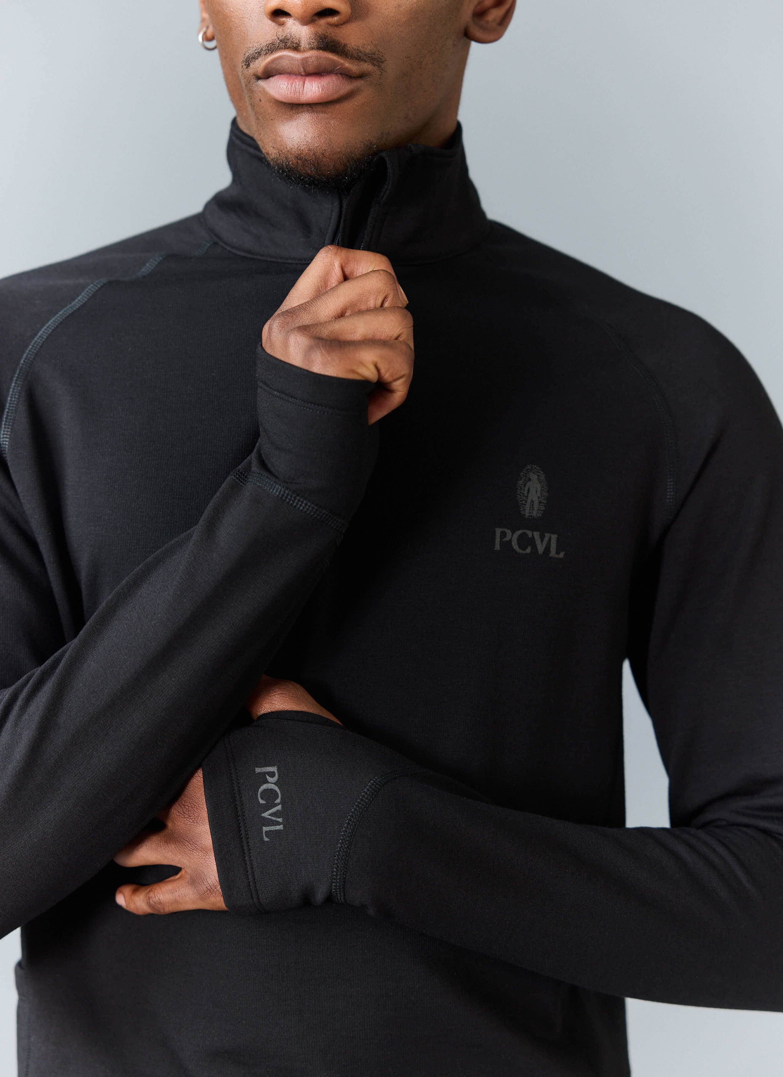 Close-up of a model wearing the black PCVL Base Layer Quarter Zip from Percival, showing the funnel neck and thumbhole cuff.