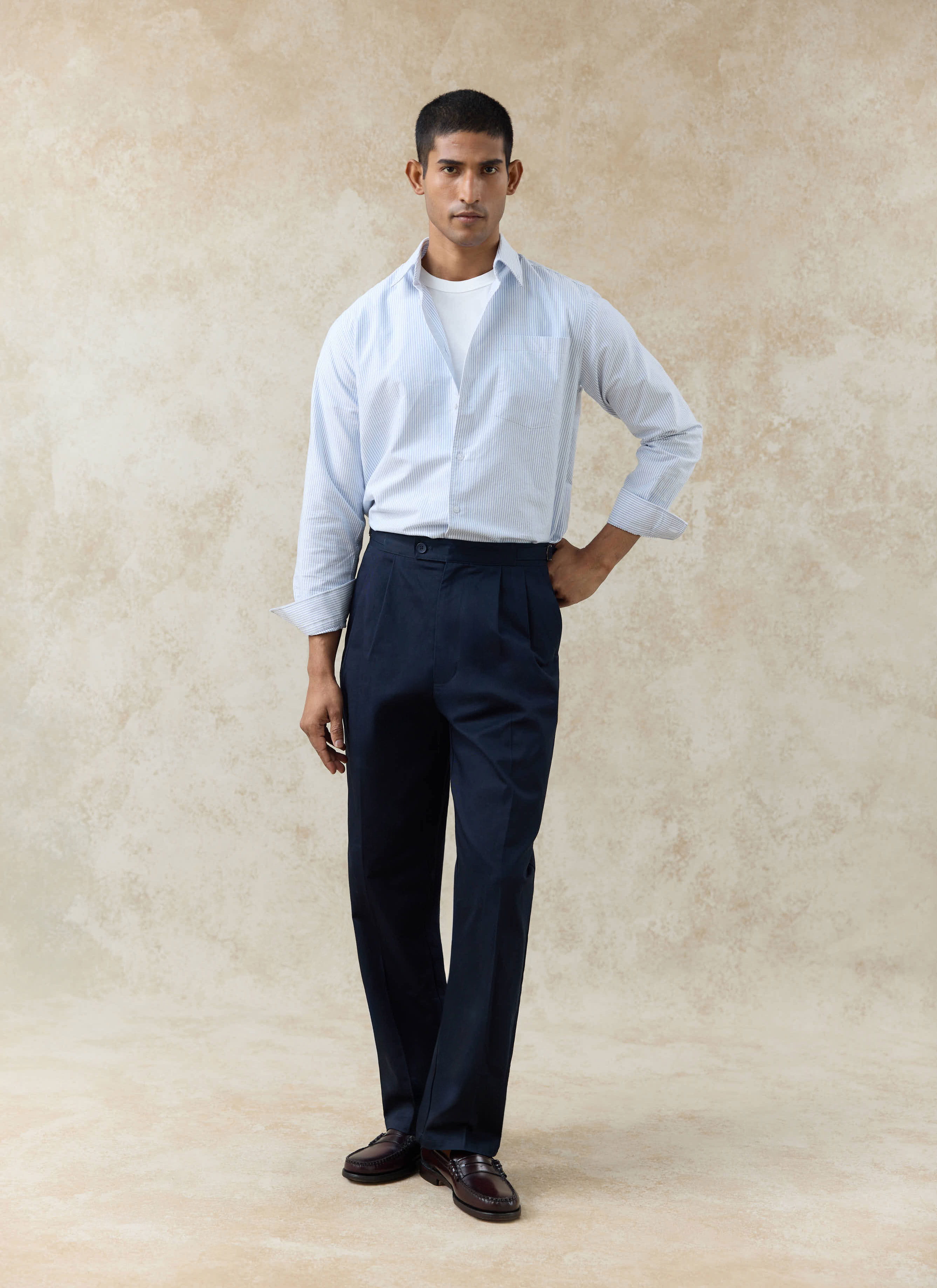 Model wears a Percival blue striped Oxford shirt and navy cotton double pleated Blake trousers against a beige wall.