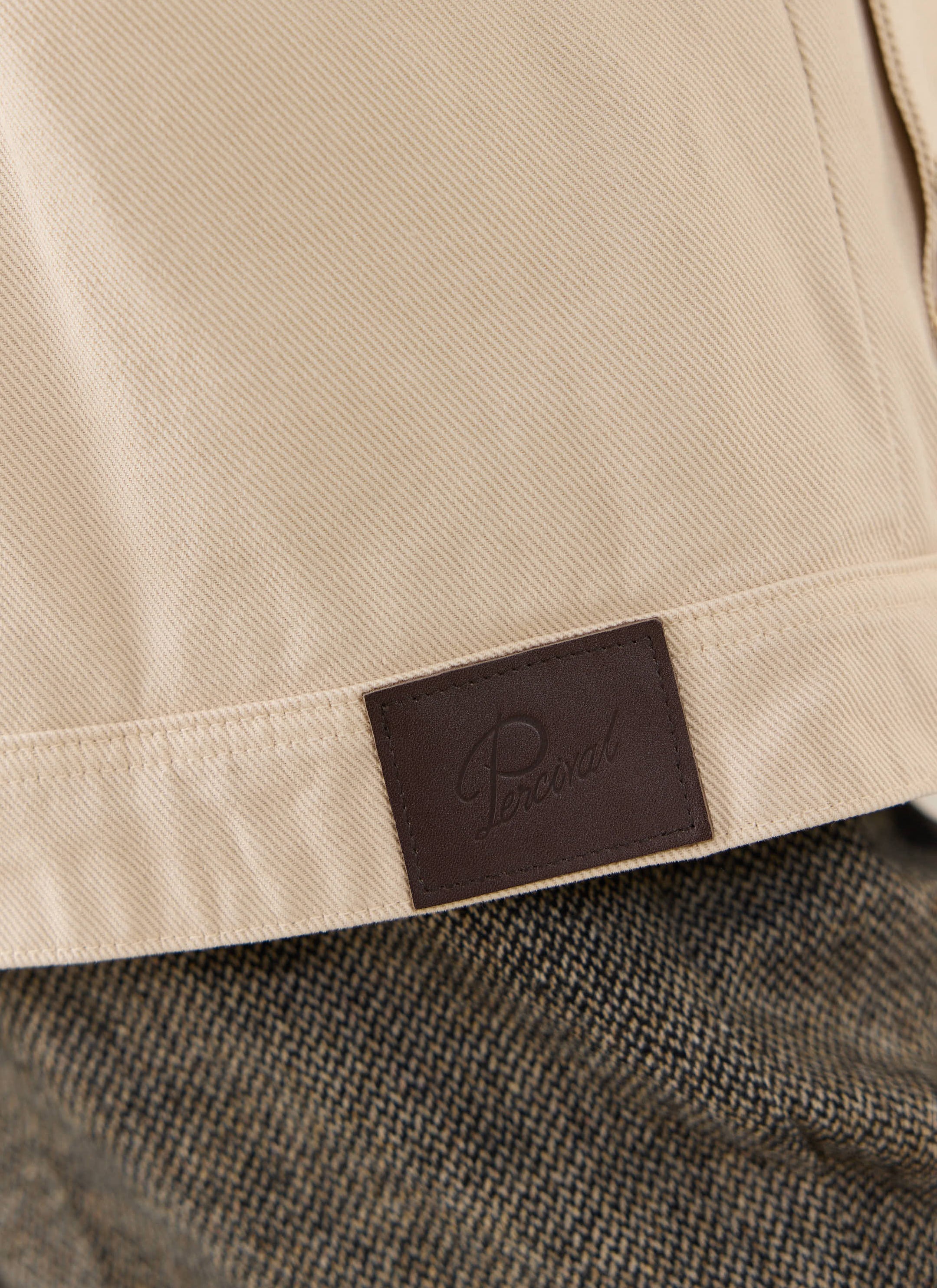 Detail of the cream cotton Percival Fontane Boxy Jacket and brown leather patch, worn over dark herringbone trousers.