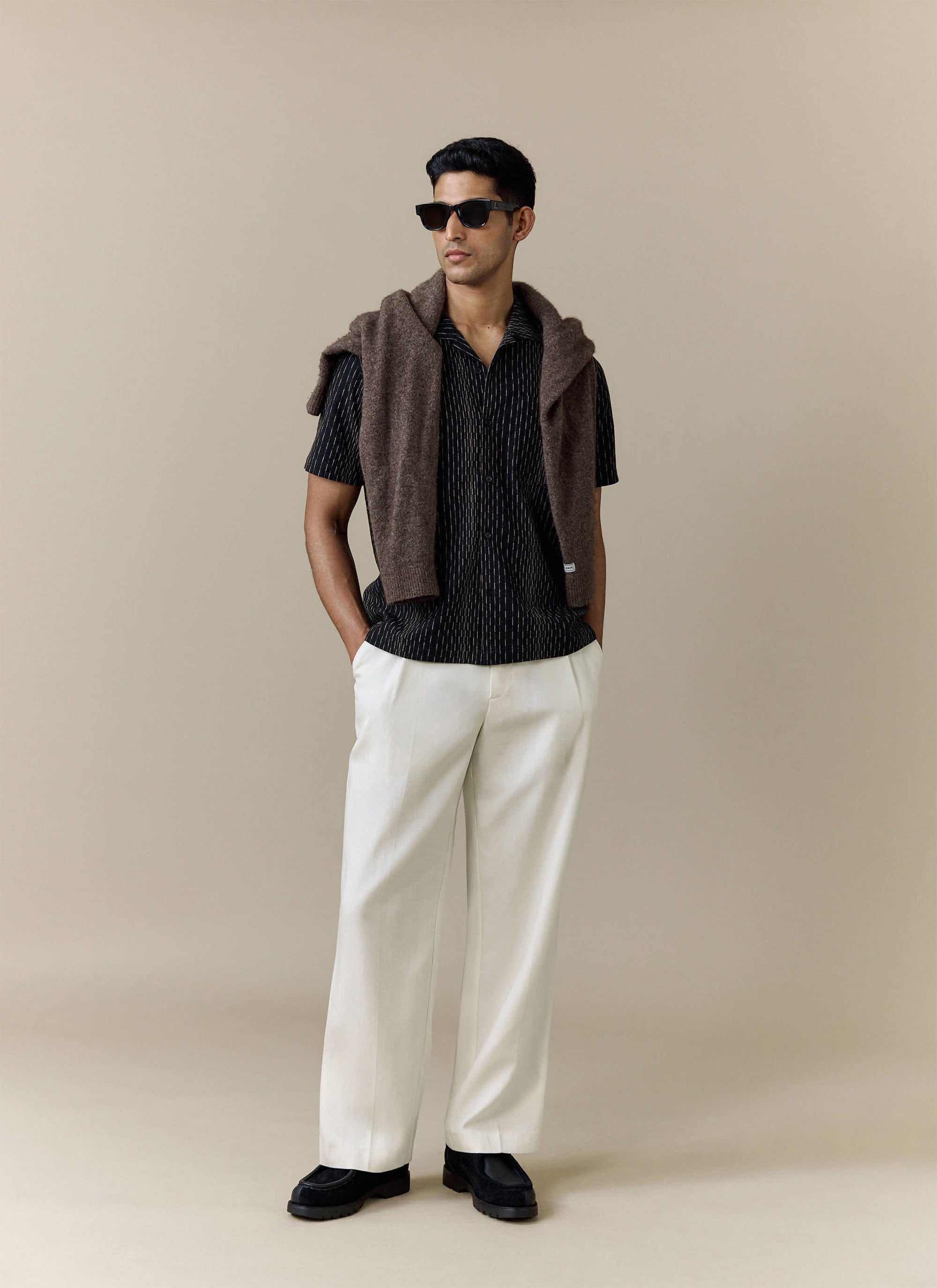 A model wears the black Percival Leland Cuban Shirt, styled with white trousers and a brown jumper over his shoulders.