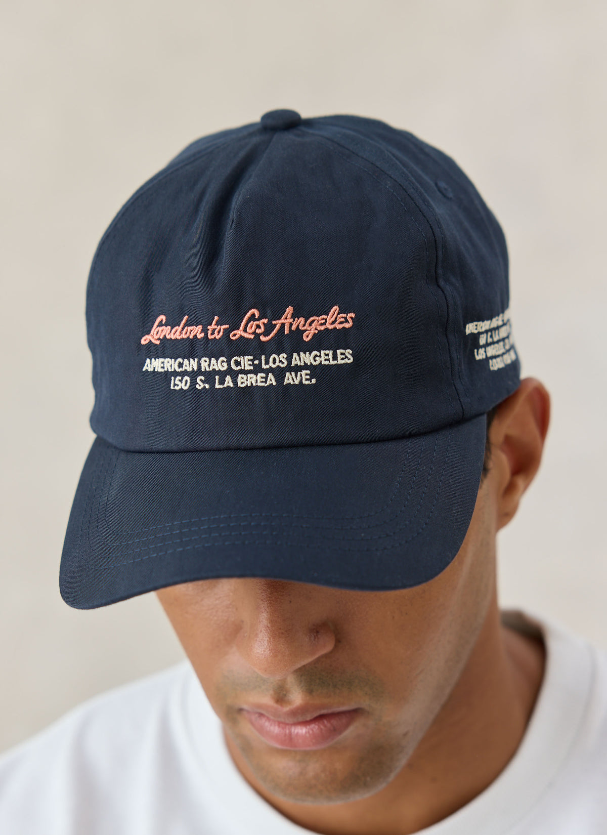A close-up of the navy blue American Rag x Percival cap featuring embroidered London to Los Angeles lettering on the front.