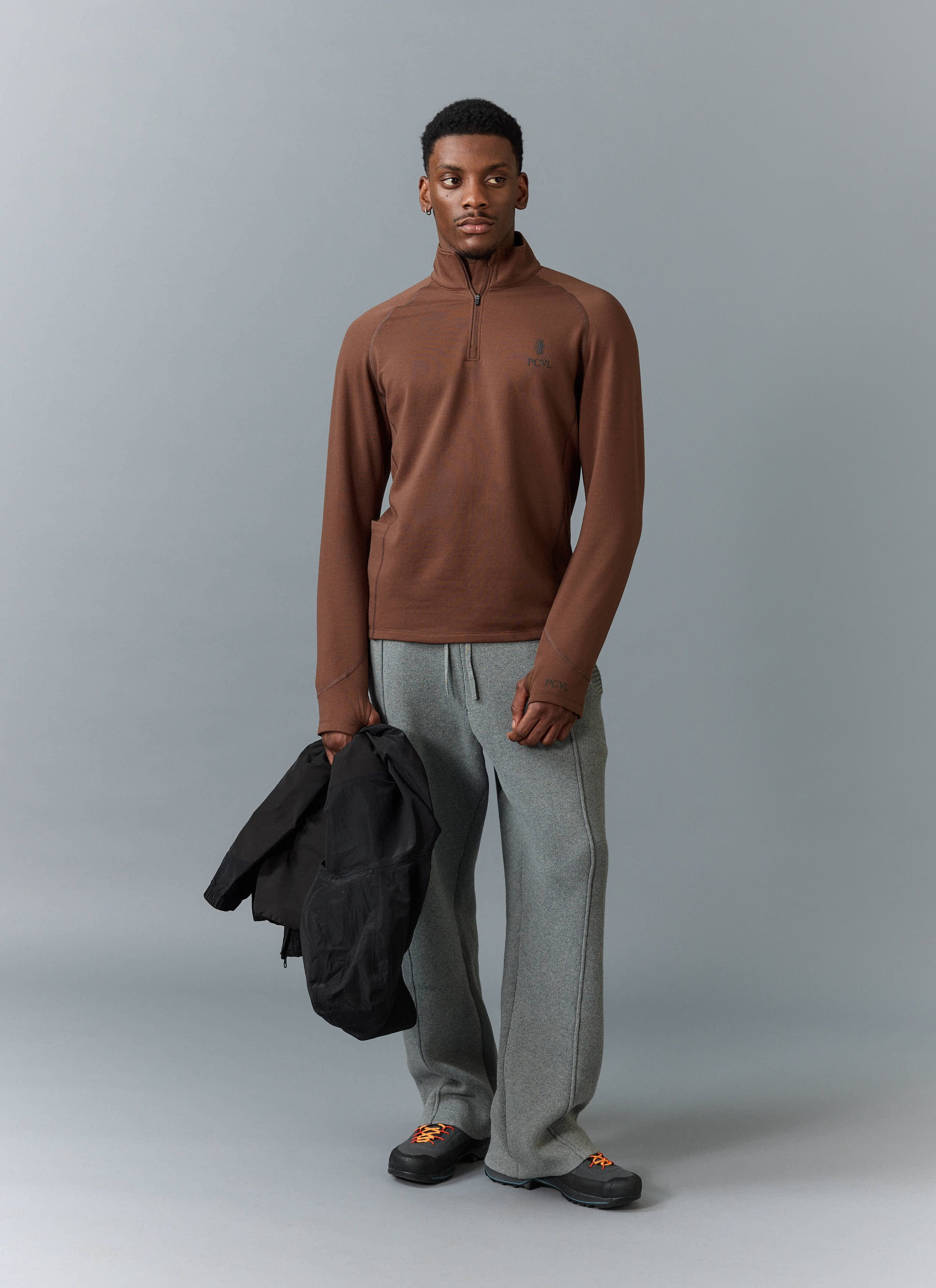 A model wears the Percival PCVL Base Layer Quarter Zip in rust, styled with grey trousers and holding a black jacket.