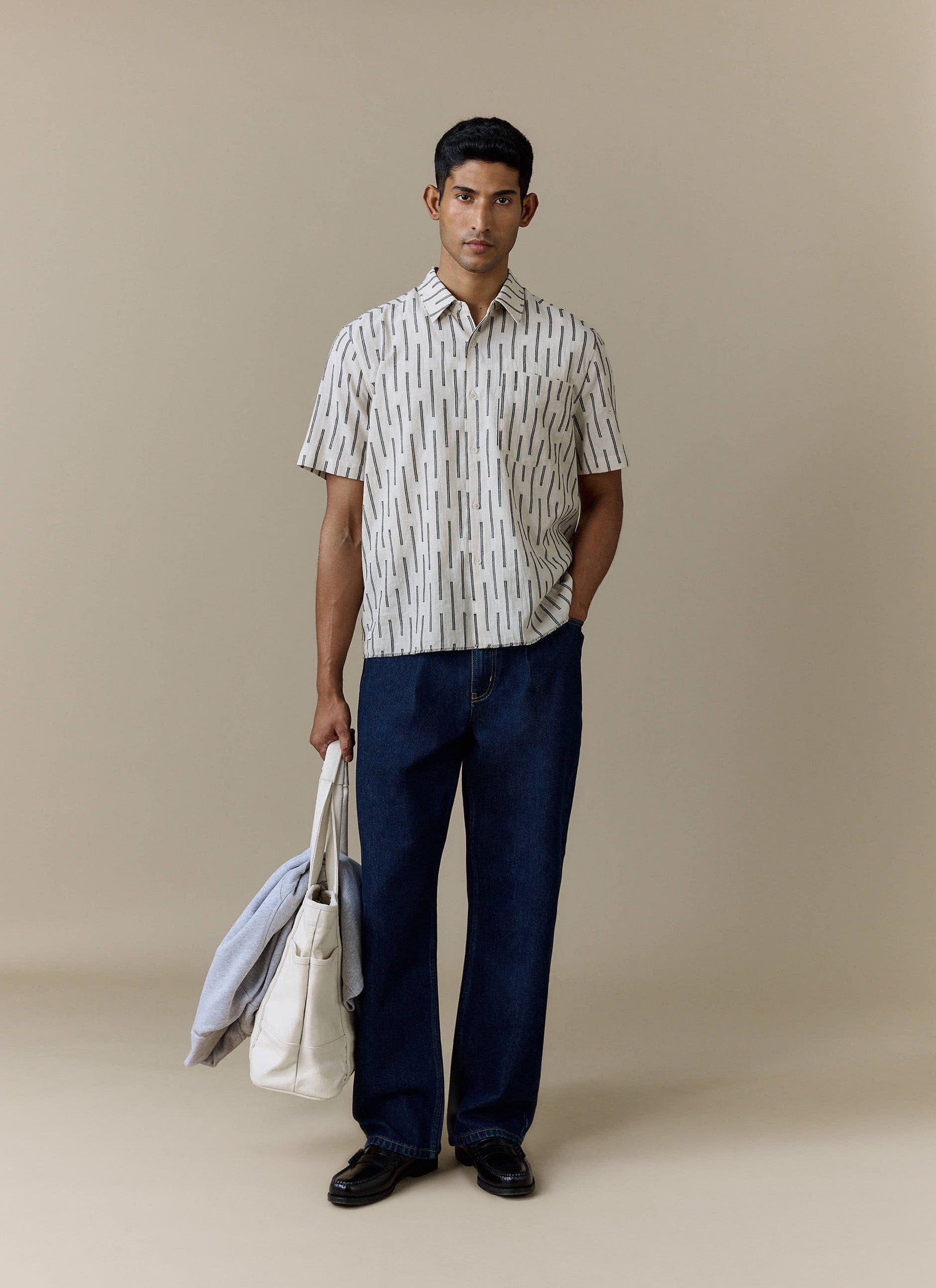 A model wears the dark indigo Pleated Denim Jeans from Percival, styled with a patterned shirt, loafers and a tote bag.