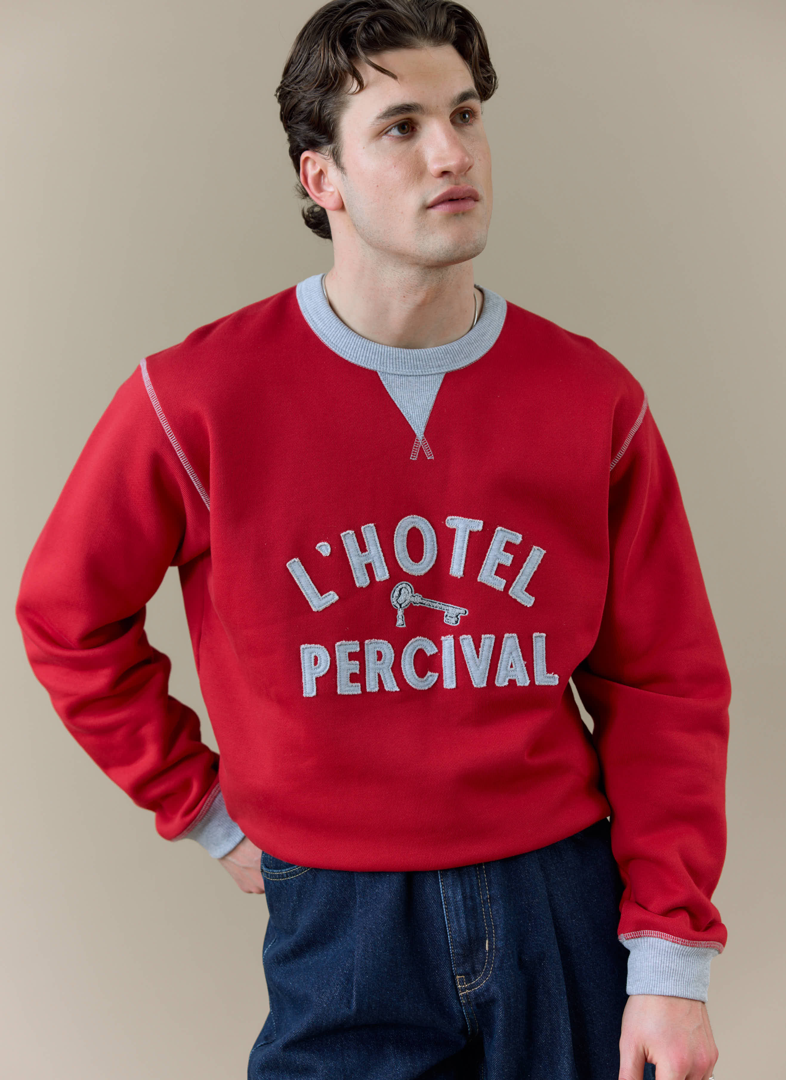 A model wearing the red Percival Ronald Sweatshirt, featuring 'L'Hotel Percival' applique and contrast grey details.