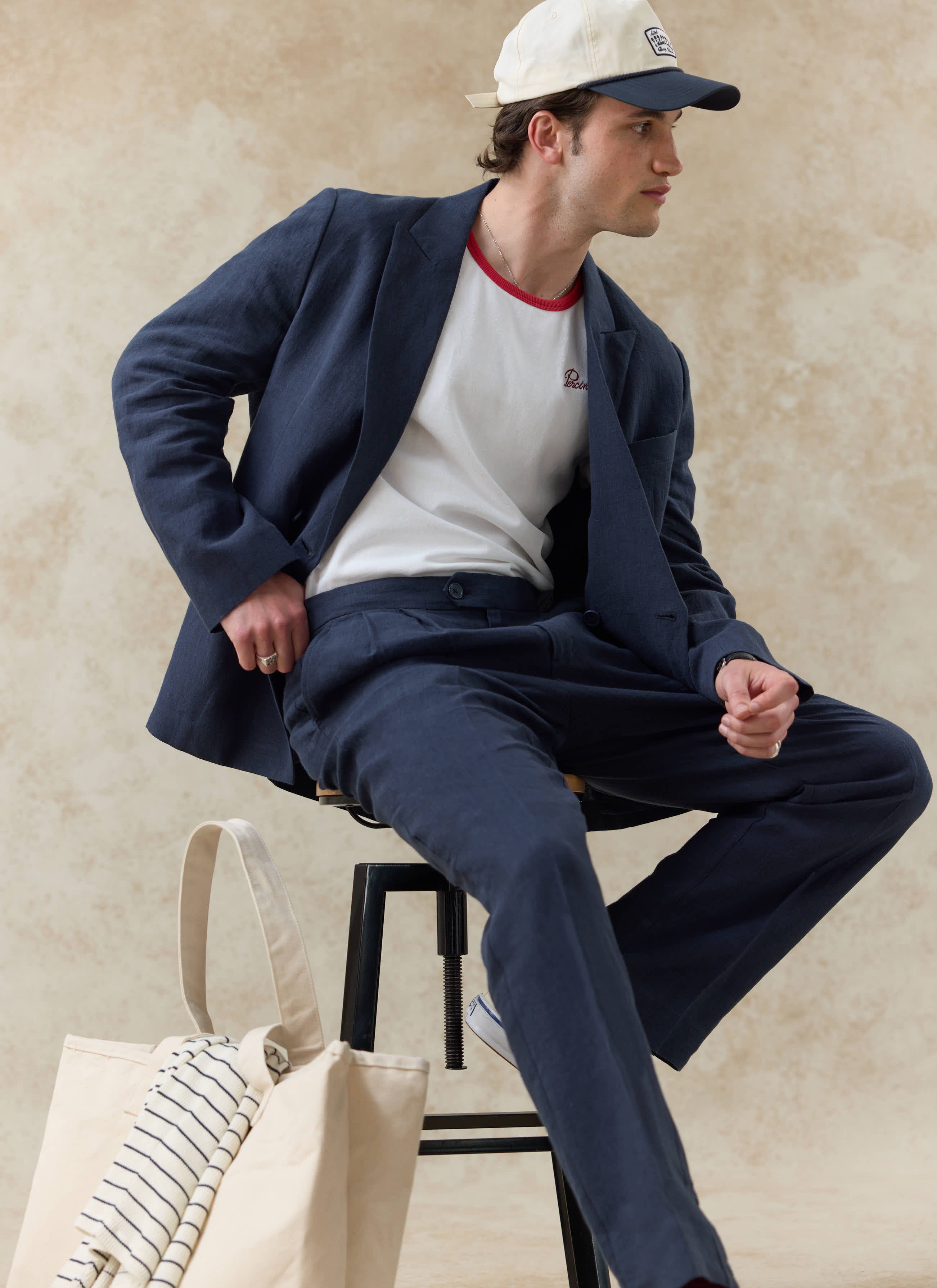 Seated model wearing the Percival Donovan Soft Trucker Cap in cream, styled with a navy linen suit and white t-shirt.