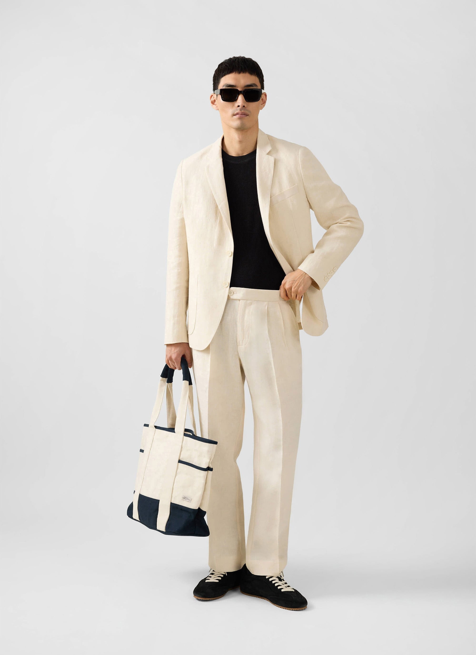 Percival cream tailored linen suit worn by a male model with sunglasses and a tote bag against a white background.
