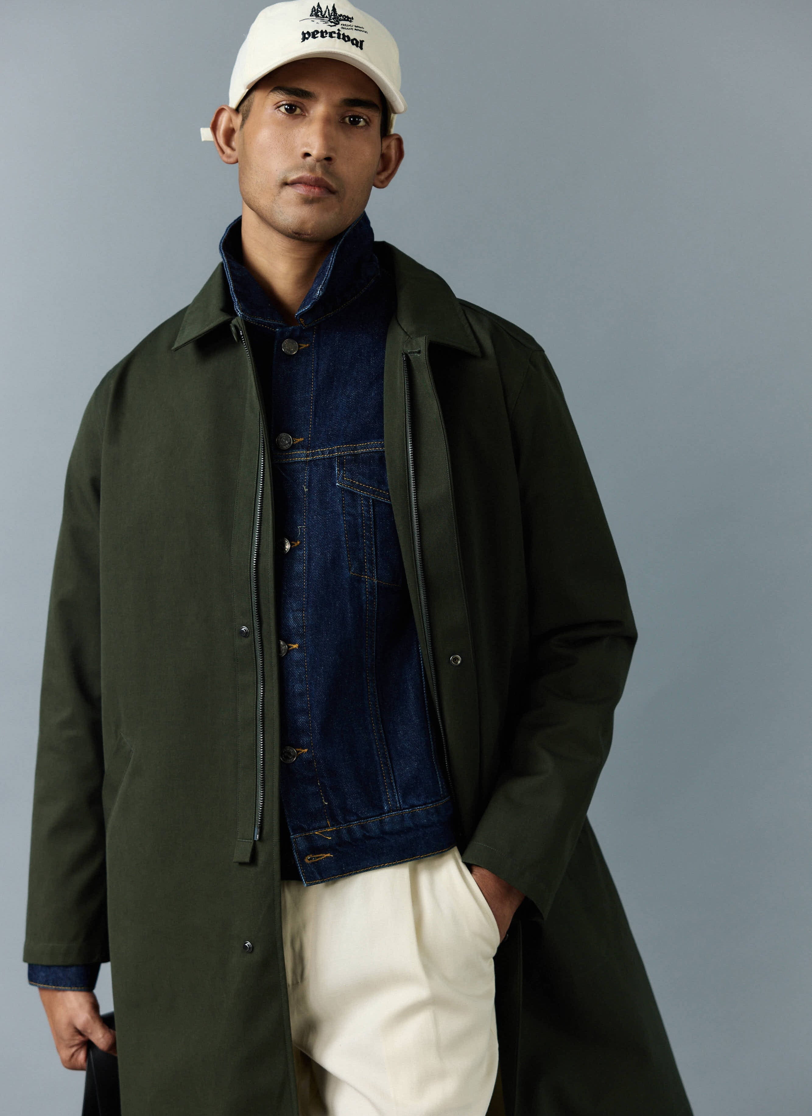 A man models the forest green Greyson Trench Coat from Percival, layered over a denim jacket and paired with a white cap.