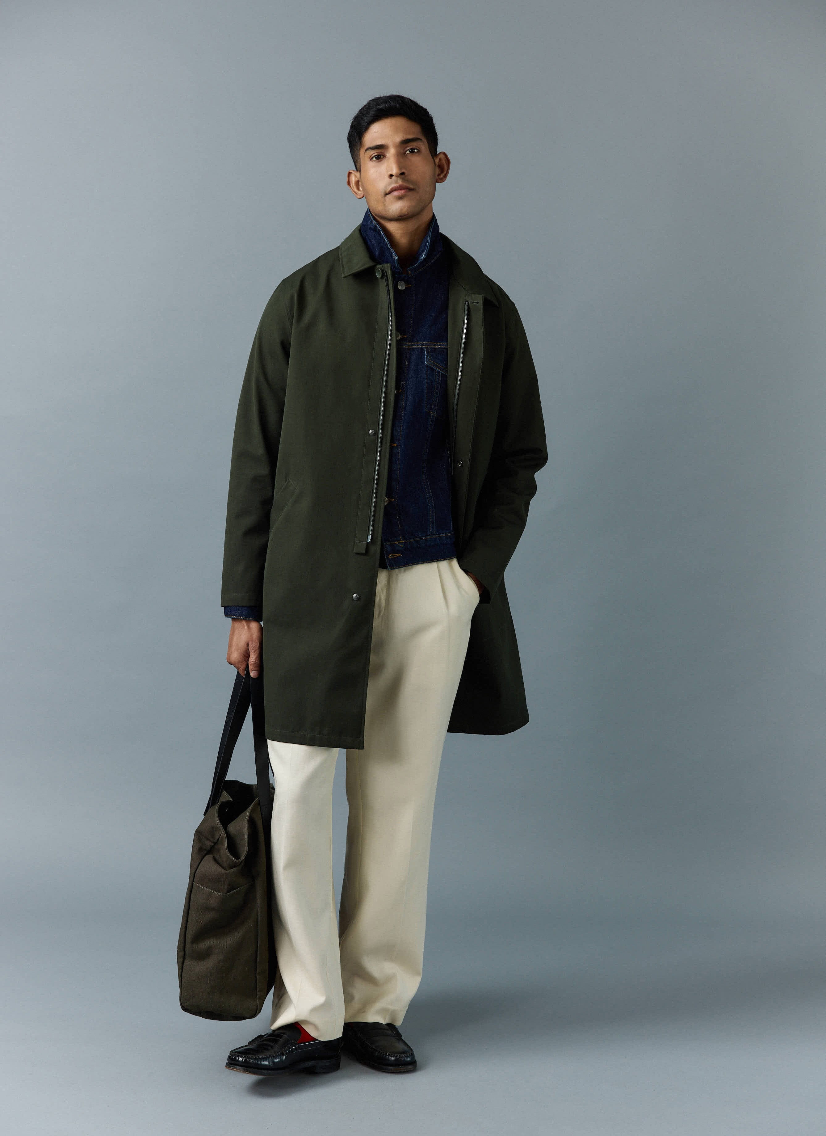 A model wears the Percival Greyson Trench Coat in Forest over a denim jacket with cream trousers and a tote bag.
