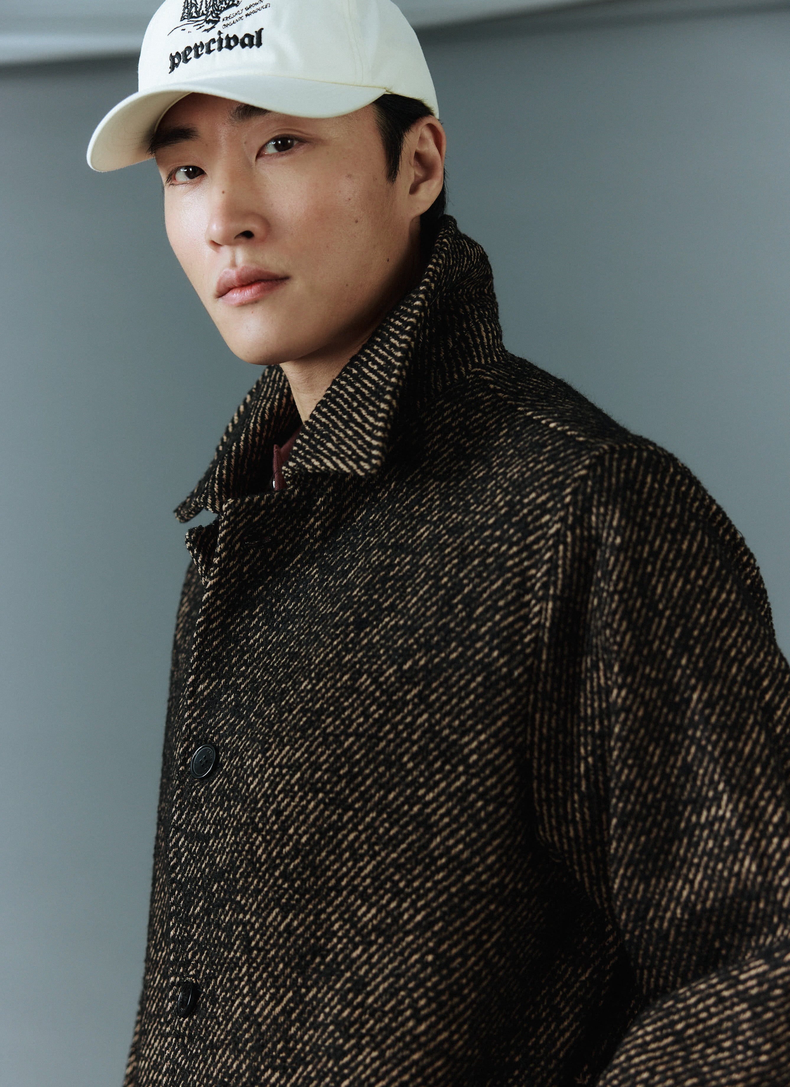 Close-up of a model wearing the Percival Minnesota Overcoat in oak twill and a white Percival baseball cap.