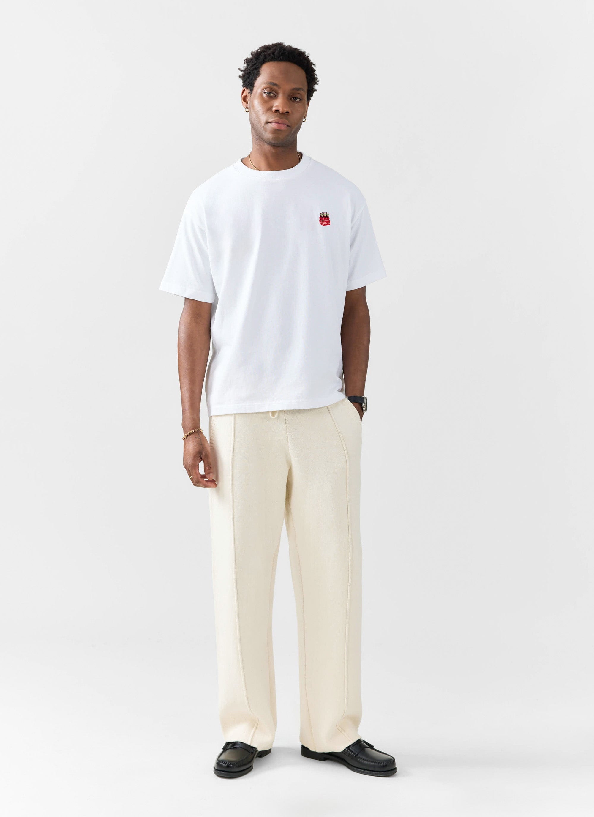 A man models the Percival Soda Bottles embroidered t-shirt in white, styled with cream trousers and black loafers.