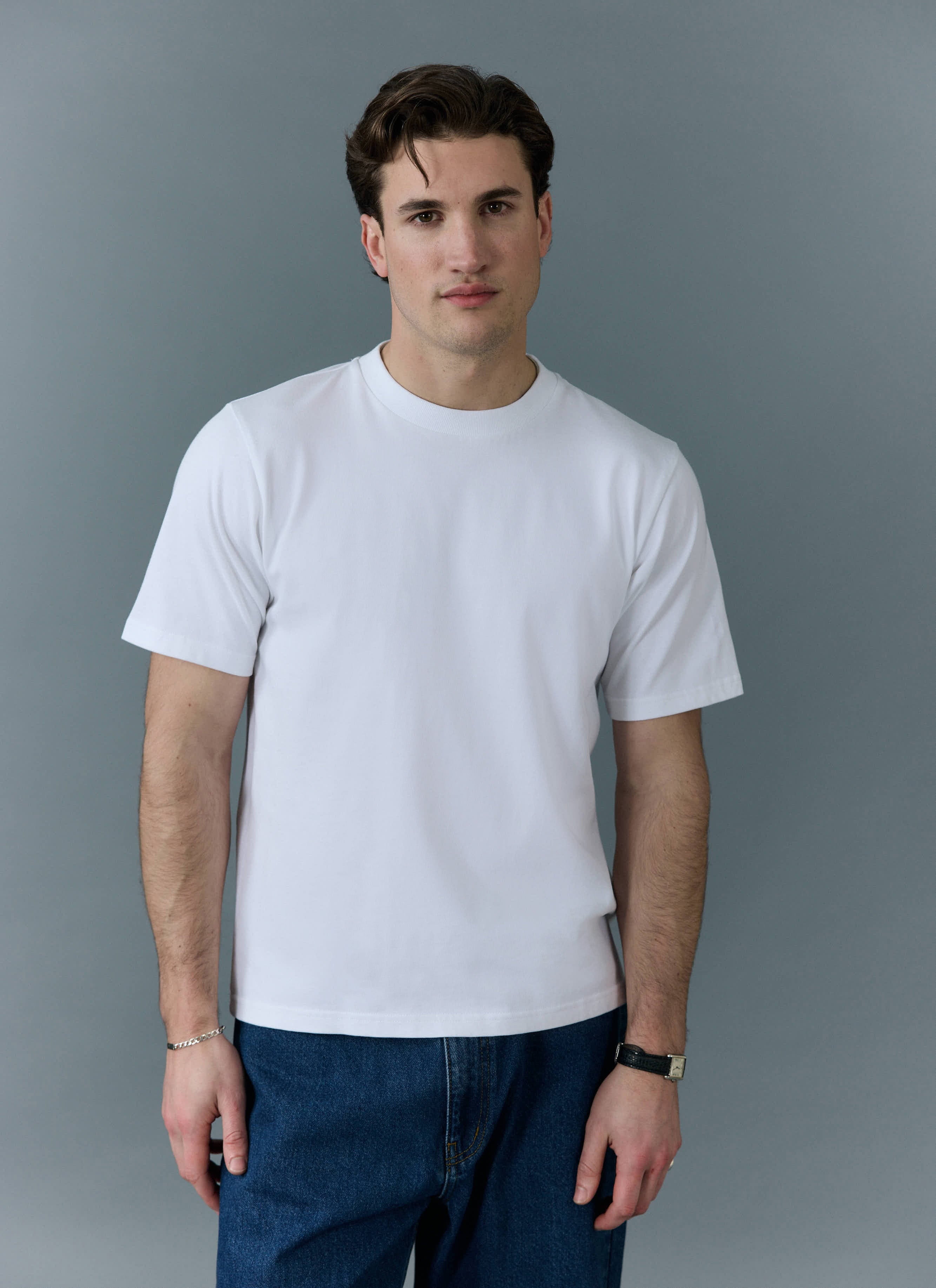 A male model wears the white Cropped Regular Fit T Shirt from Percival with blue jeans against a grey studio background.