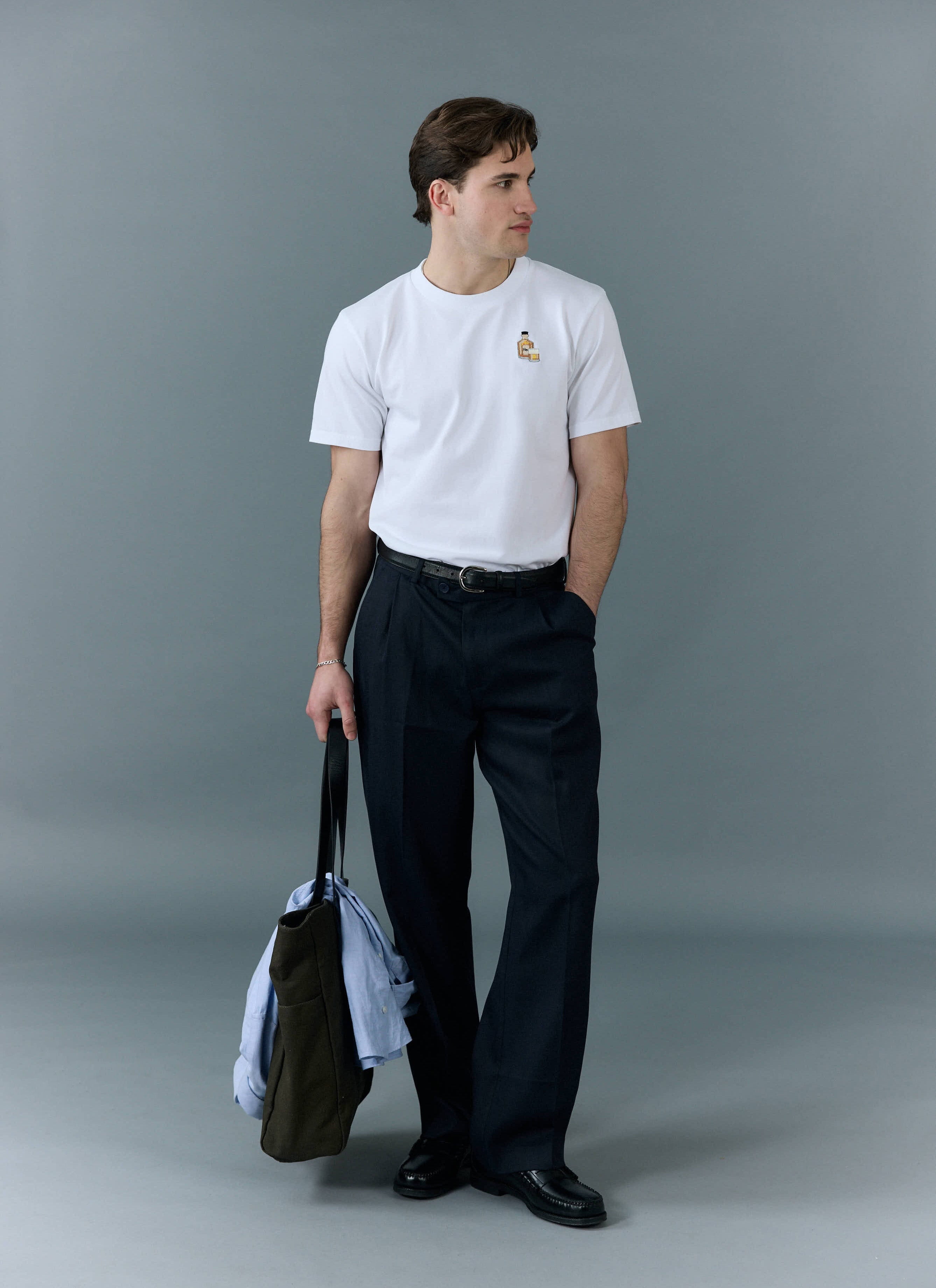 A man models the white Percival Old Fashioned T Shirt, styled with smart dark trousers and carrying a tote bag.