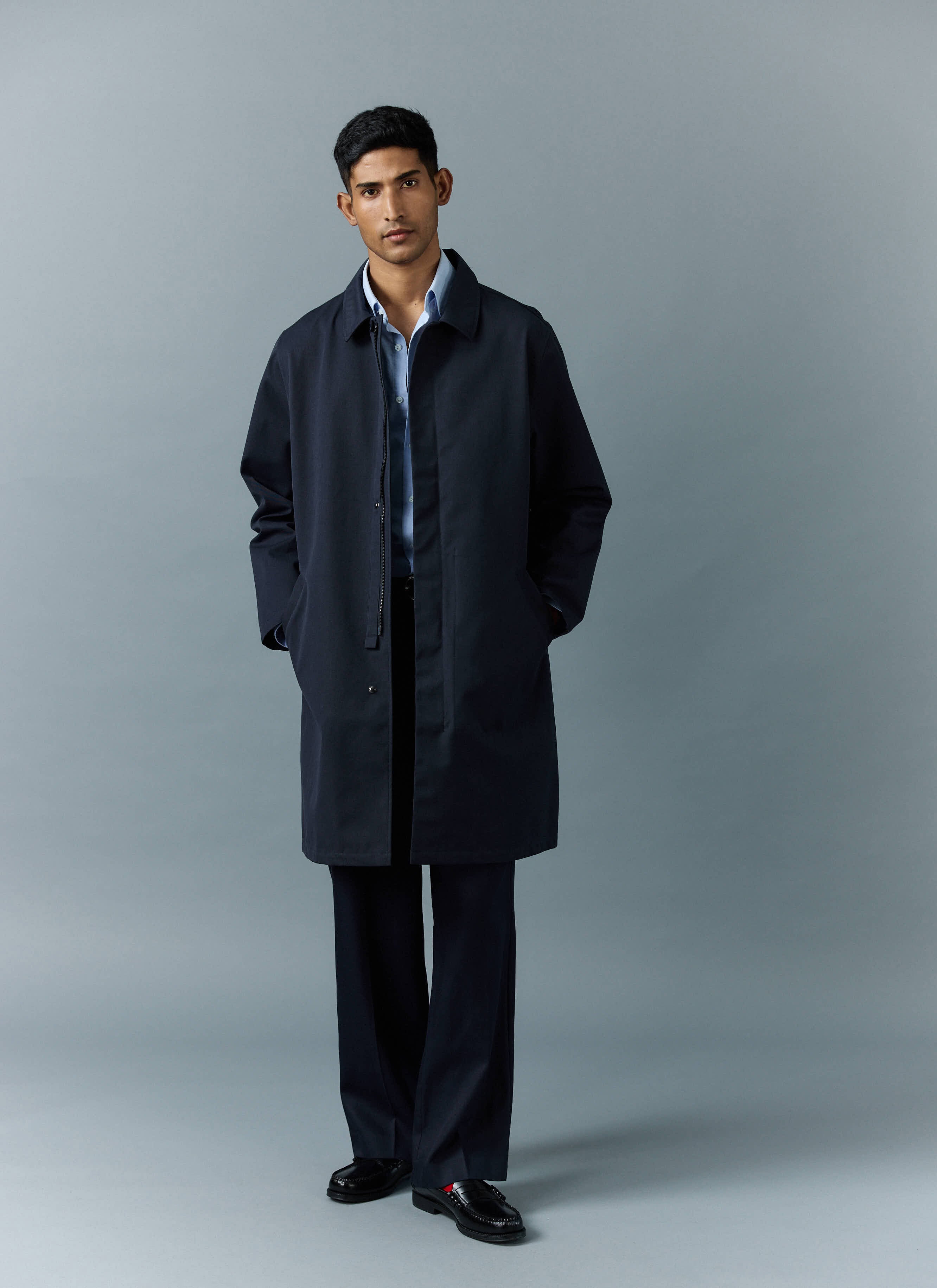 A model wears the Percival Greyson Long Trench Coat in navy, styled over a light blue shirt and dark trousers.