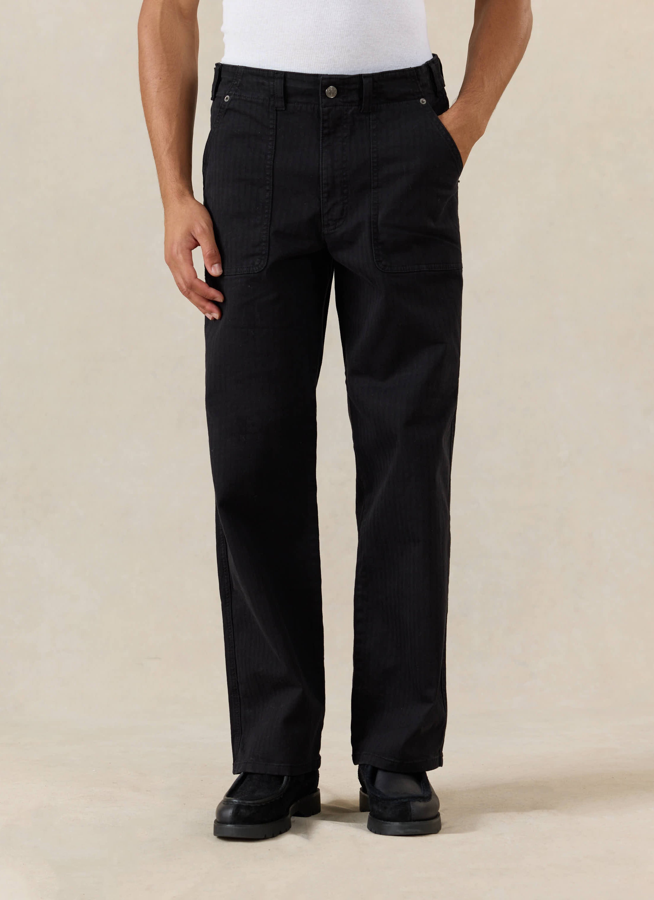 A model wears the Percival black cotton Lungo Carpenter Trousers, featuring utility patch pockets and a straight-leg fit.
