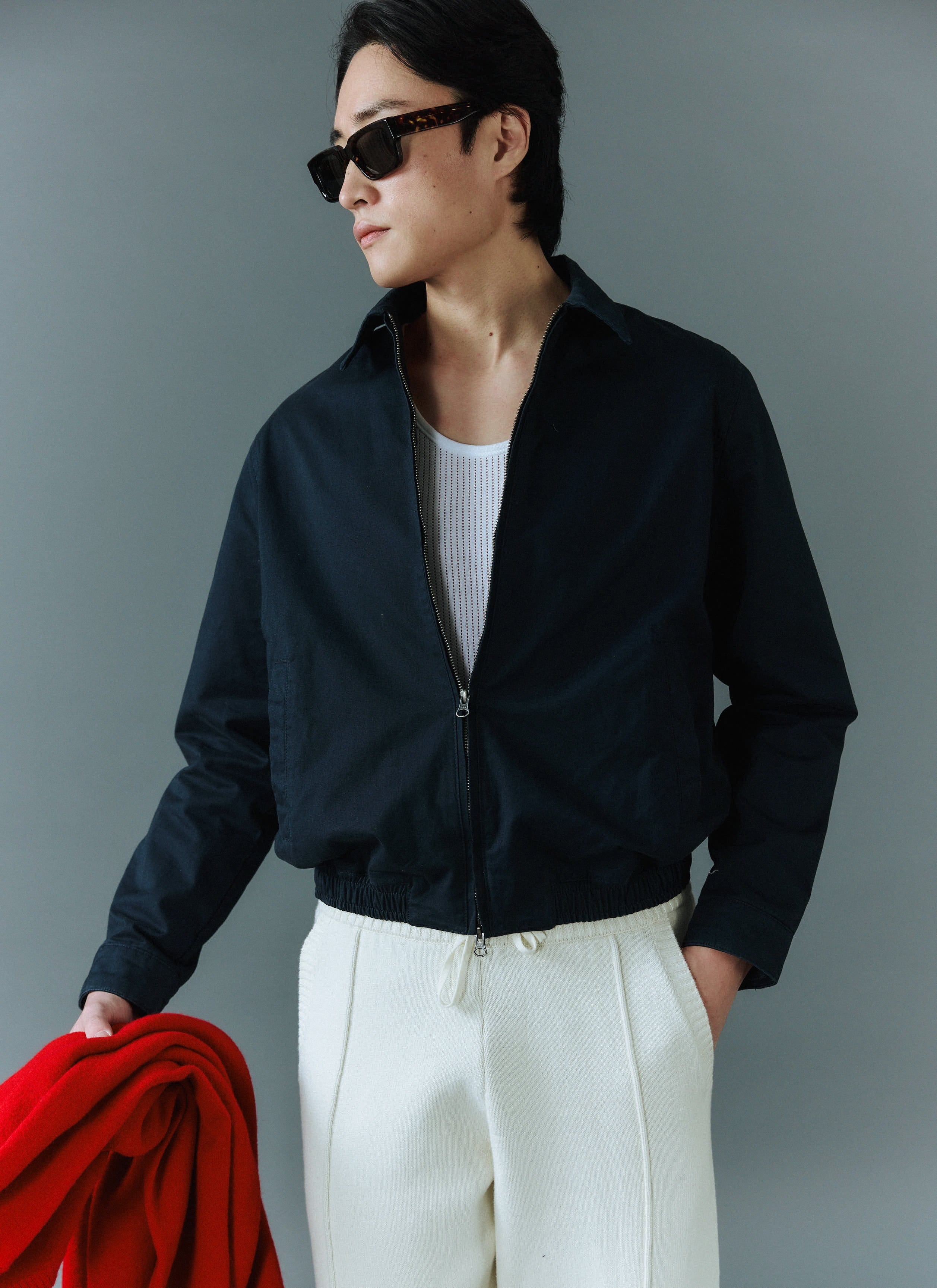 A man models the Percival Leo Harrington Jacket in navy cotton, styled with a white vest, cream trousers and sunglasses.