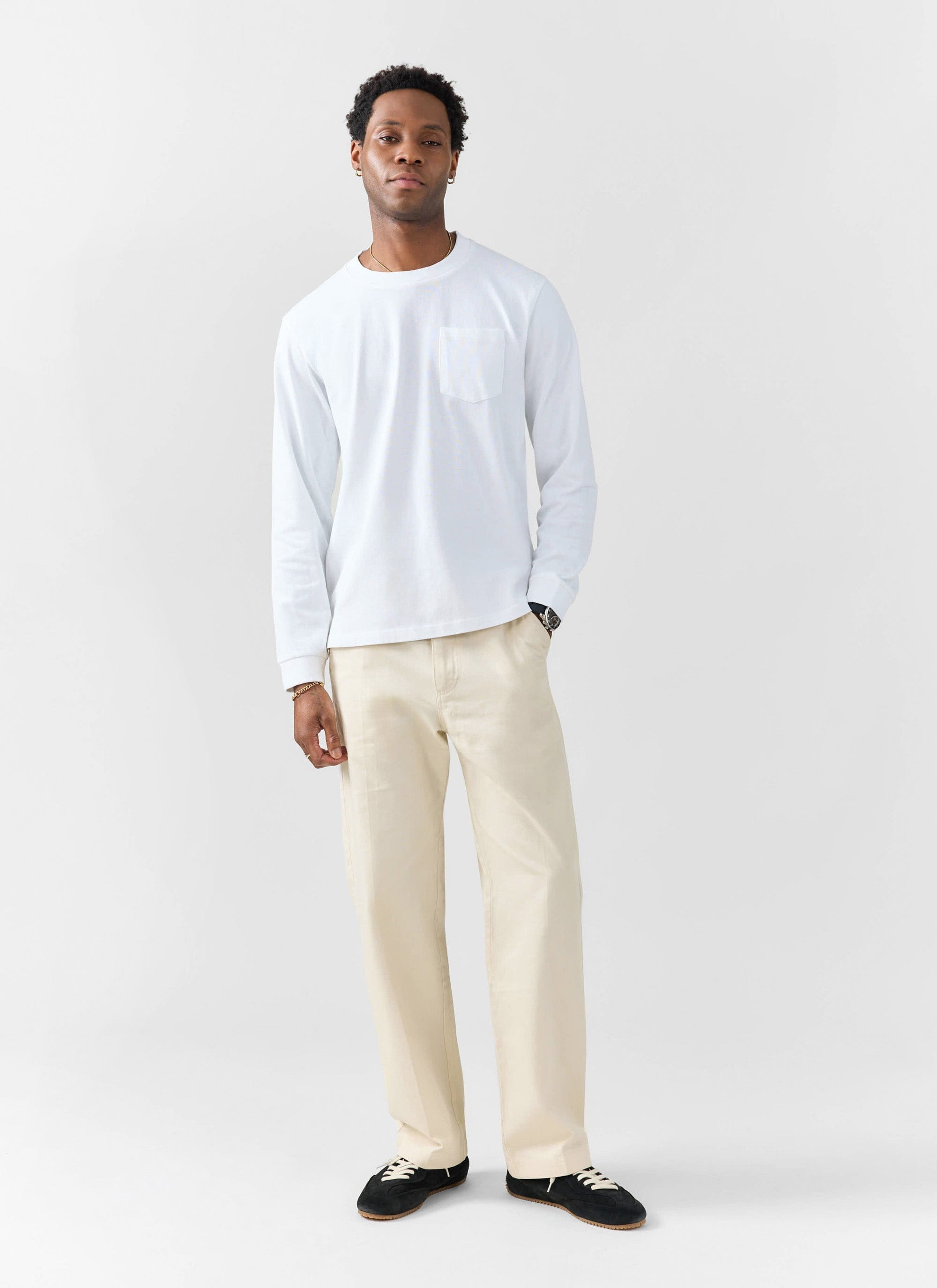 A man models the Percival Long Sleeve Pocket T-Shirt in white, paired with cream wide-leg trousers against white background.