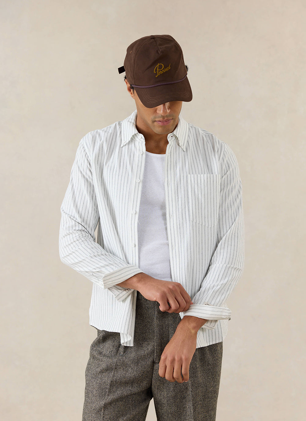 A man wears the white Percival Fabian Striped Shirt in cotton, paired with a brown cap and textured trousers.