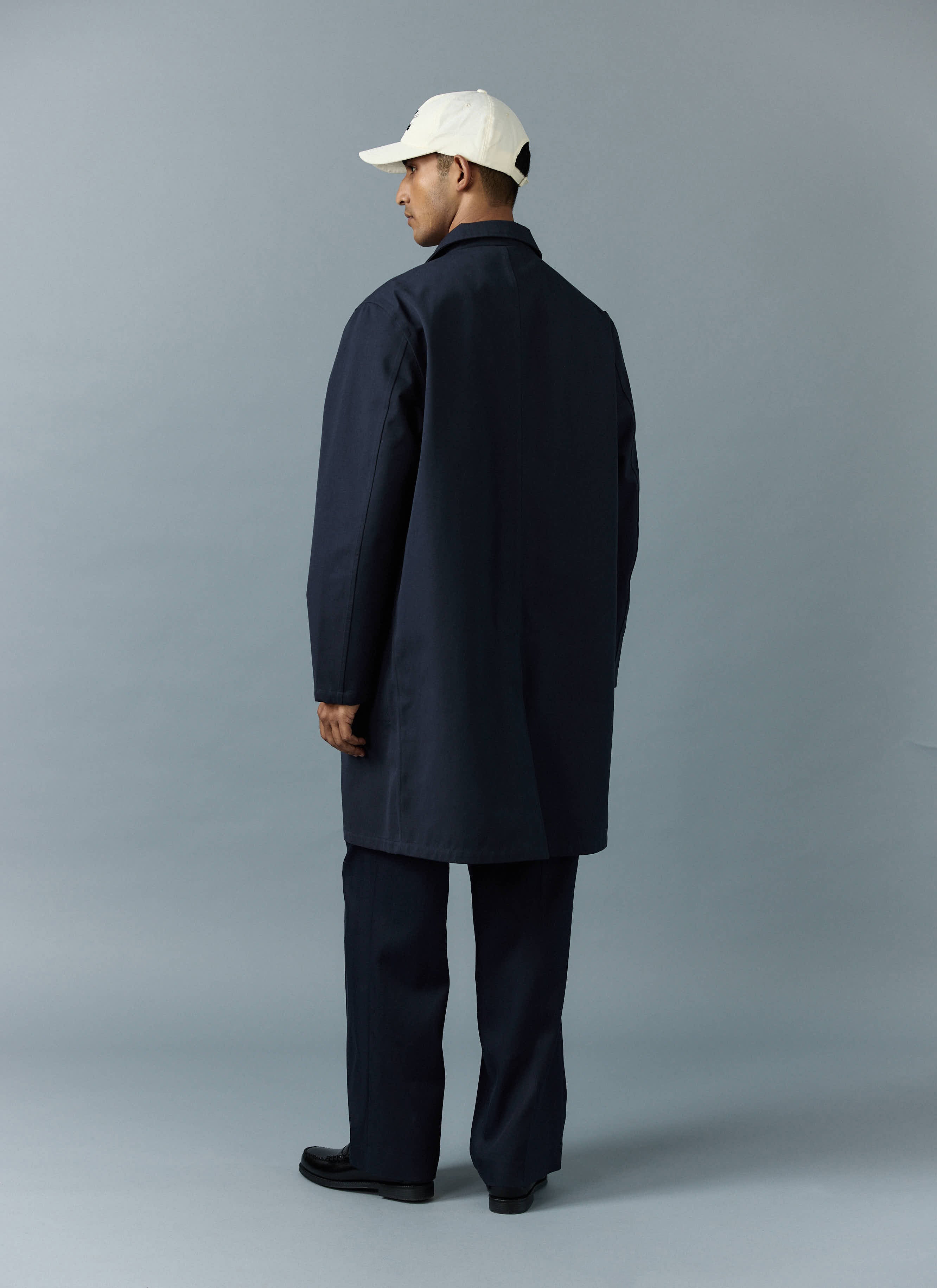 A model wears the Percival Greyson Long Trench Coat in navy, seen from behind against a grey studio background.