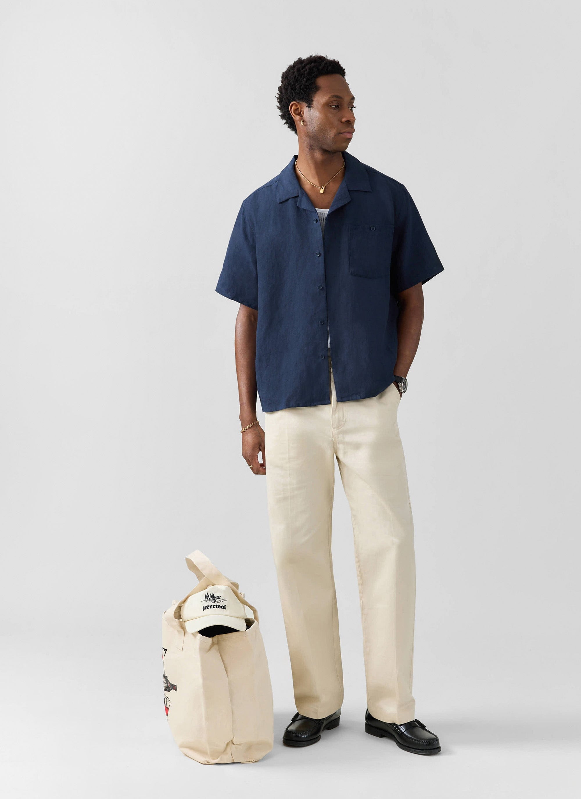A man models the Percival navy linen short sleeve Cuban shirt, with cream trousers and a Percival tote bag at his feet.