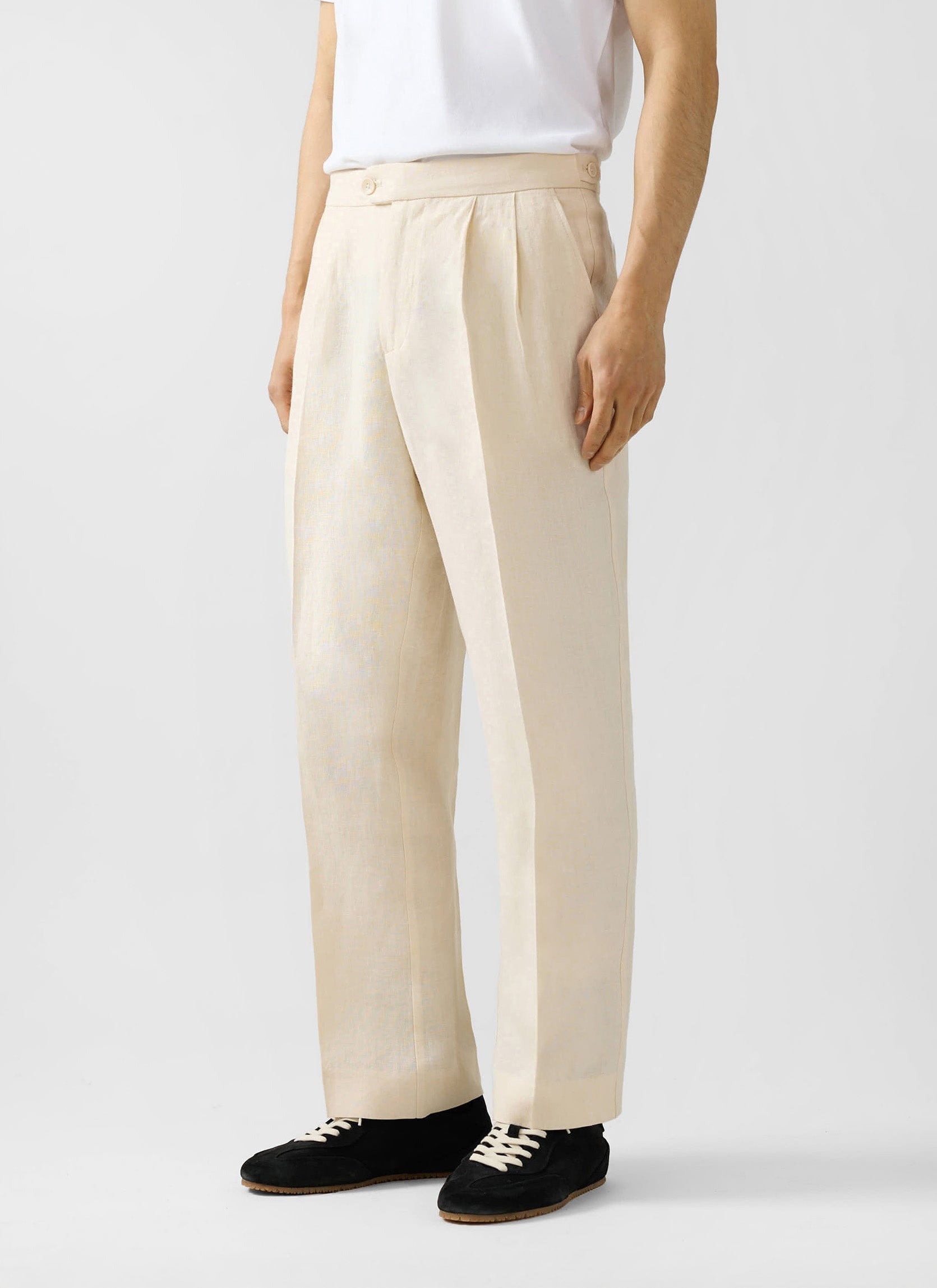 A model stands wearing the Percival Pleated Tailored Trousers in cream linen with a white t-shirt and black trainers.