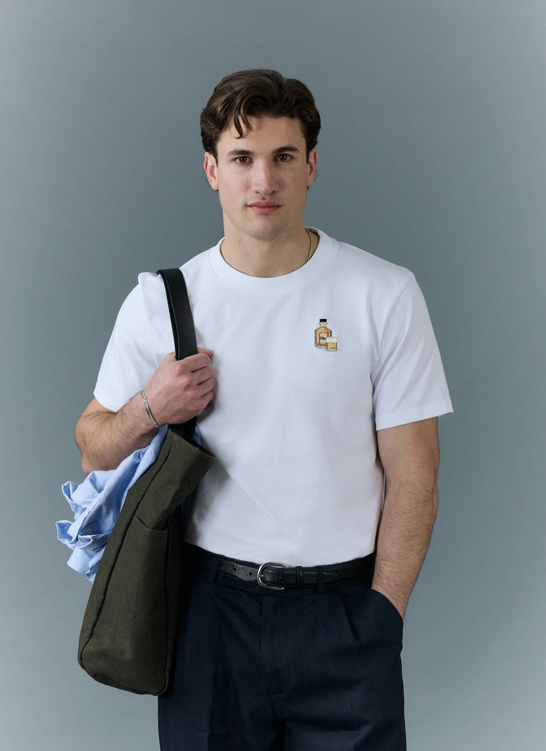 A model wears the white Old Fashioned T-shirt from Percival, paired with dark trousers and carrying a tote bag.