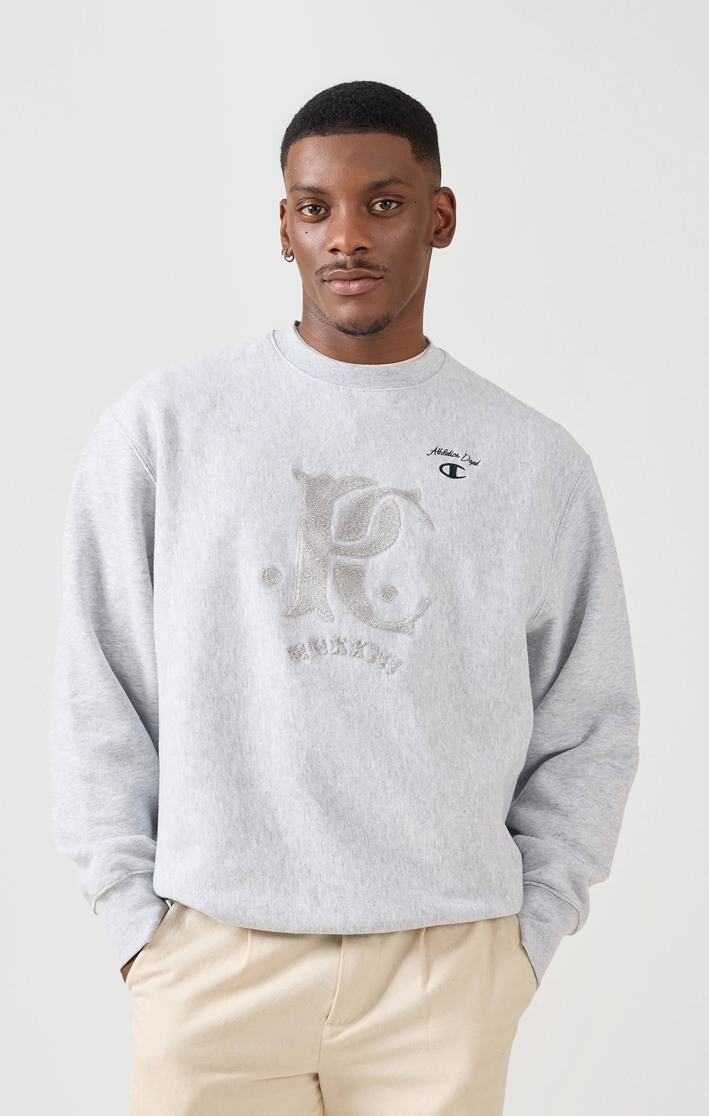 A model wears the grey Chenille Patch Sweatshirt from the Champion x Percival collaboration against a white background.