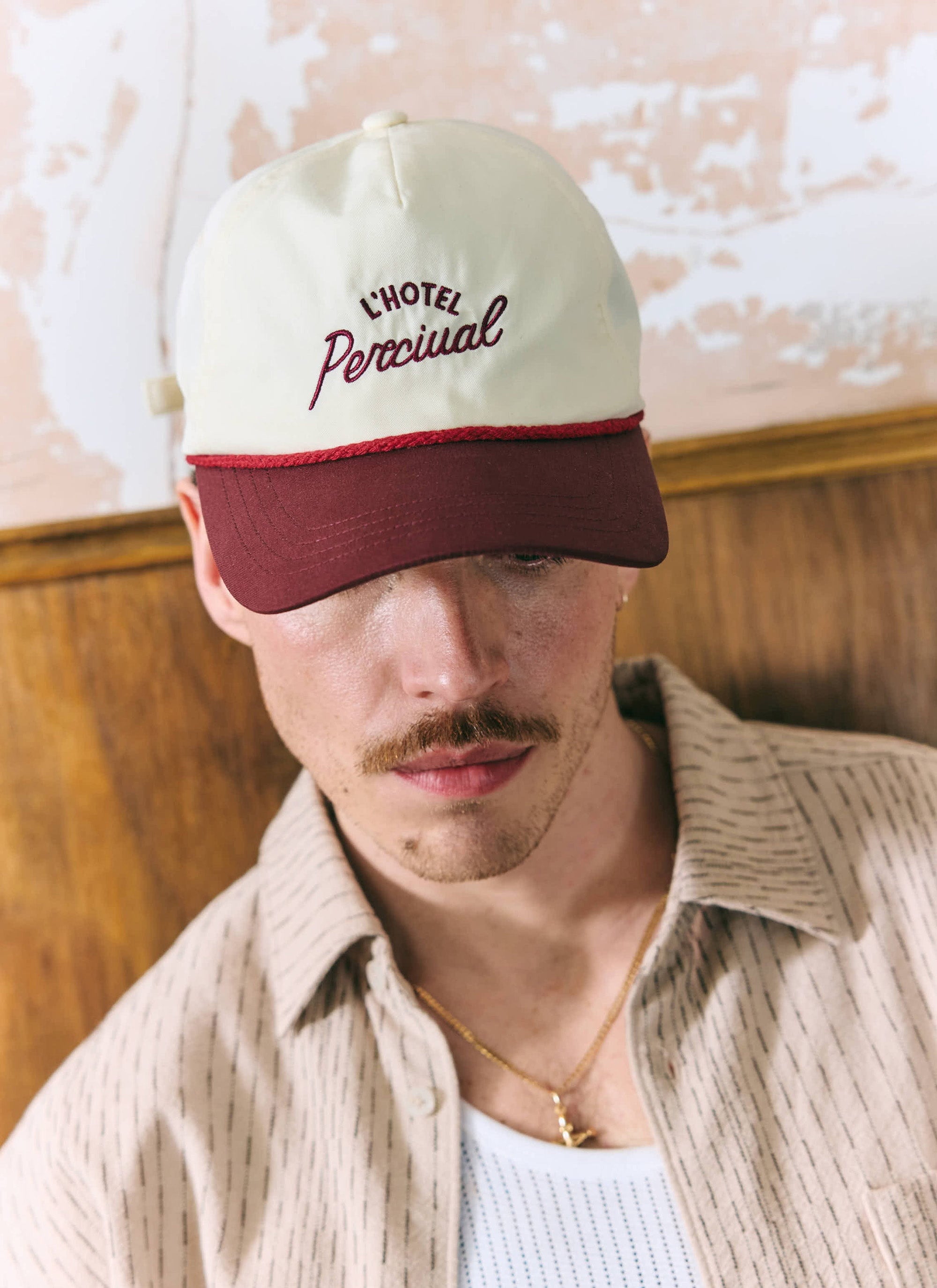 A close-up of a model wearing the ecru and burgundy L'Hotel Percival Soft Trucker Cap.