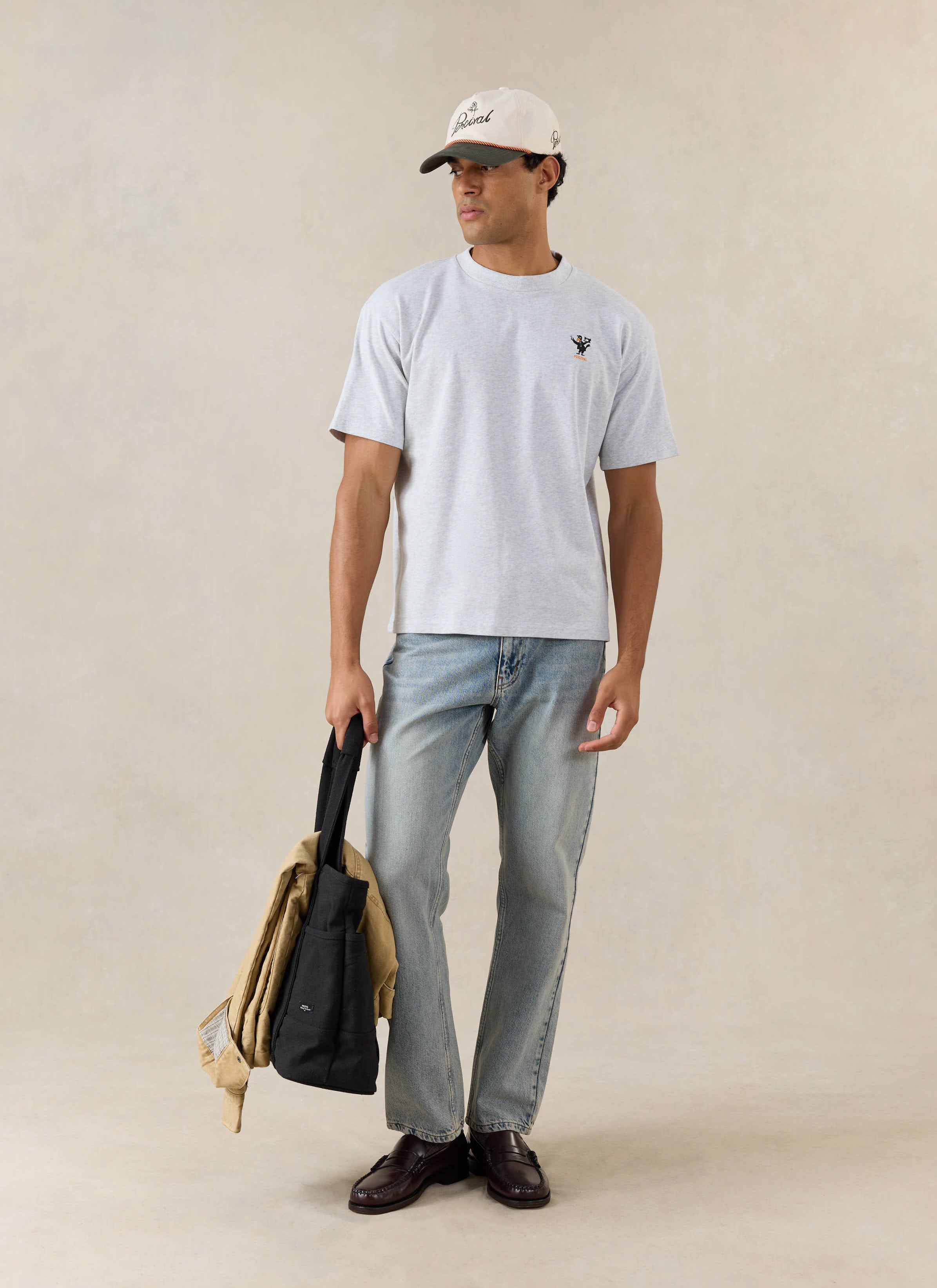 A model wears the athletic grey Percival Atticus t-shirt with light blue jeans, cap, and tote bag in a studio.