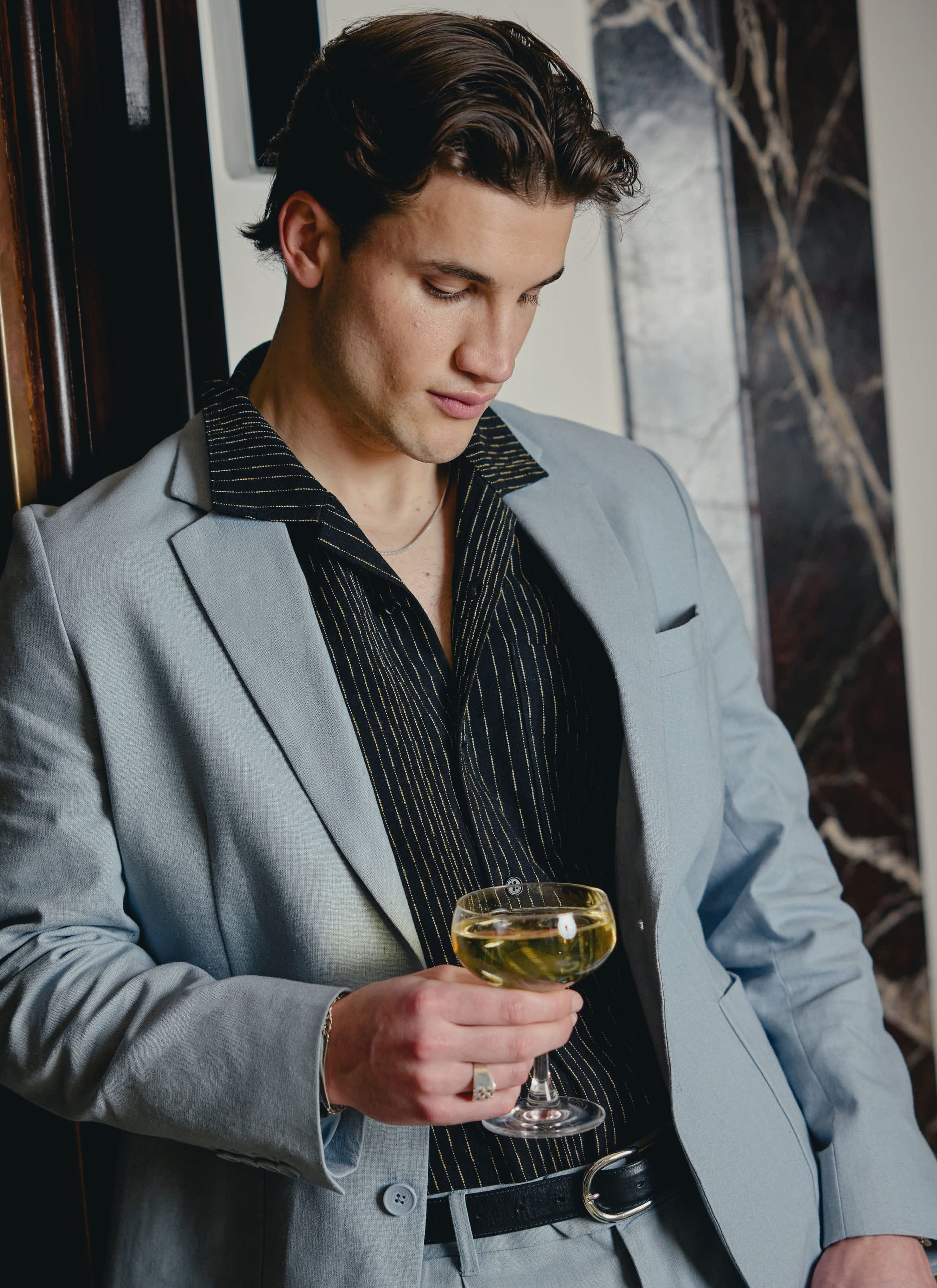 A model wears the Percival Tailored Linen Blazer in light blue with a black striped shirt, holding a glass of champagne.