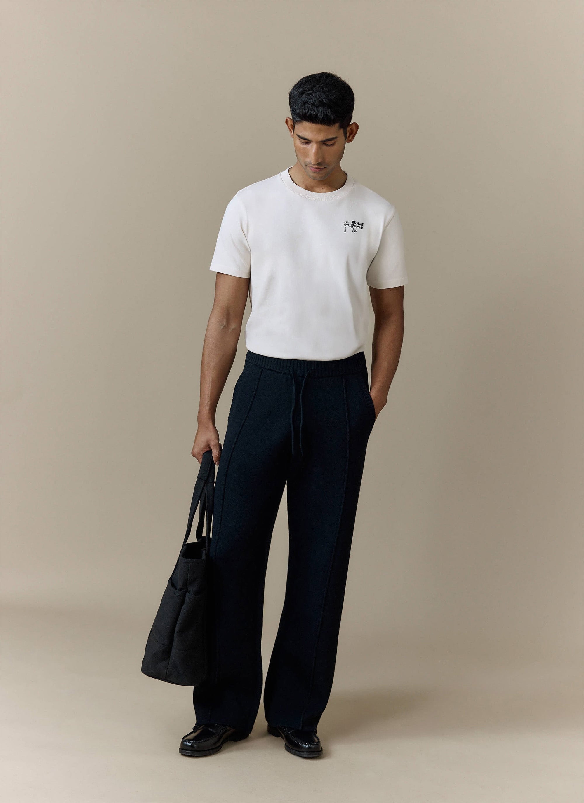 A model wears the Percival Chronicle Knitted Straight Leg Trackpants in black, styled with a white t-shirt and black loafers.