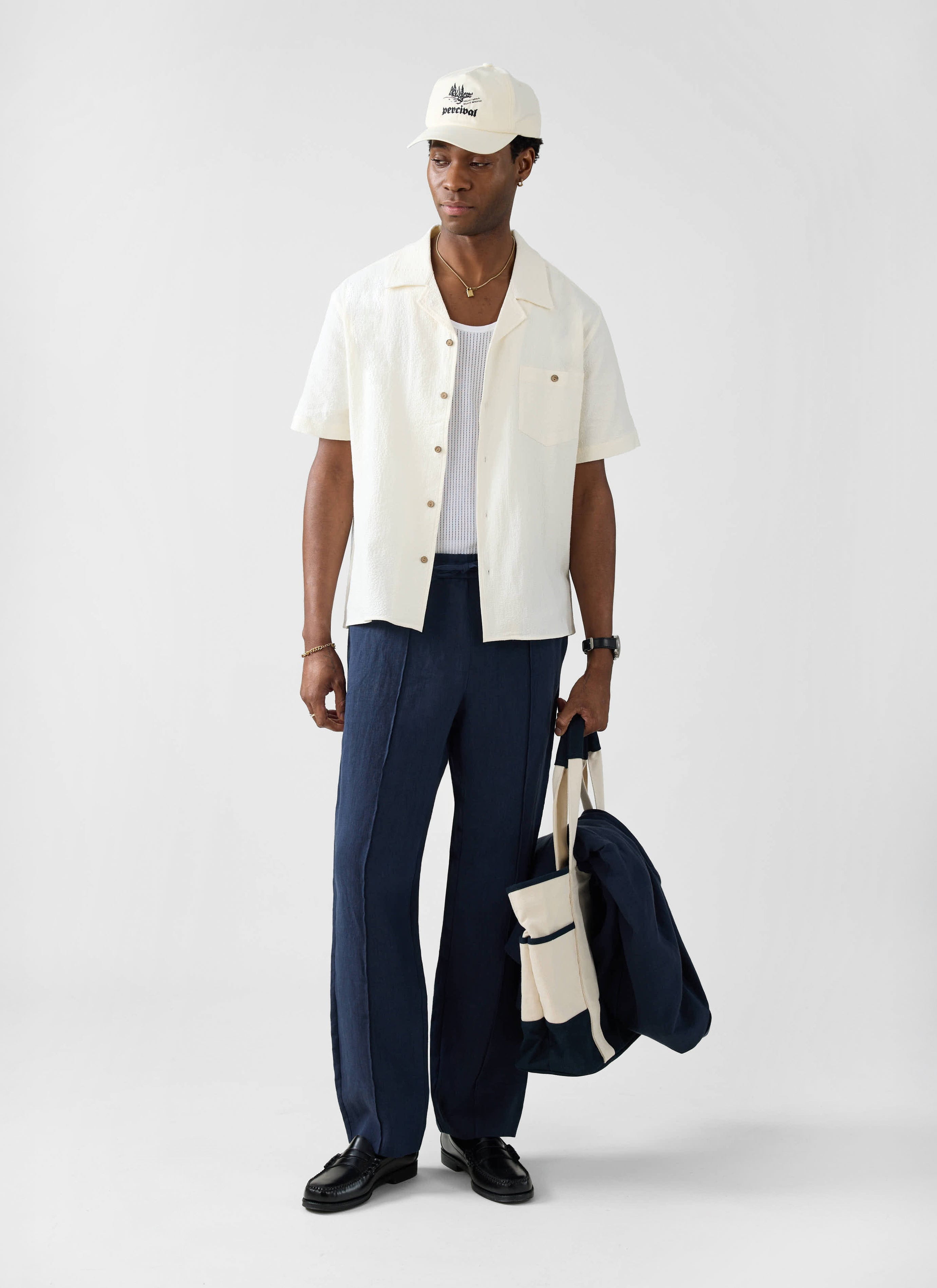 Model wears Percival ecru seersucker cuban shirt with navy trousers, black loafers and a Percival cap.