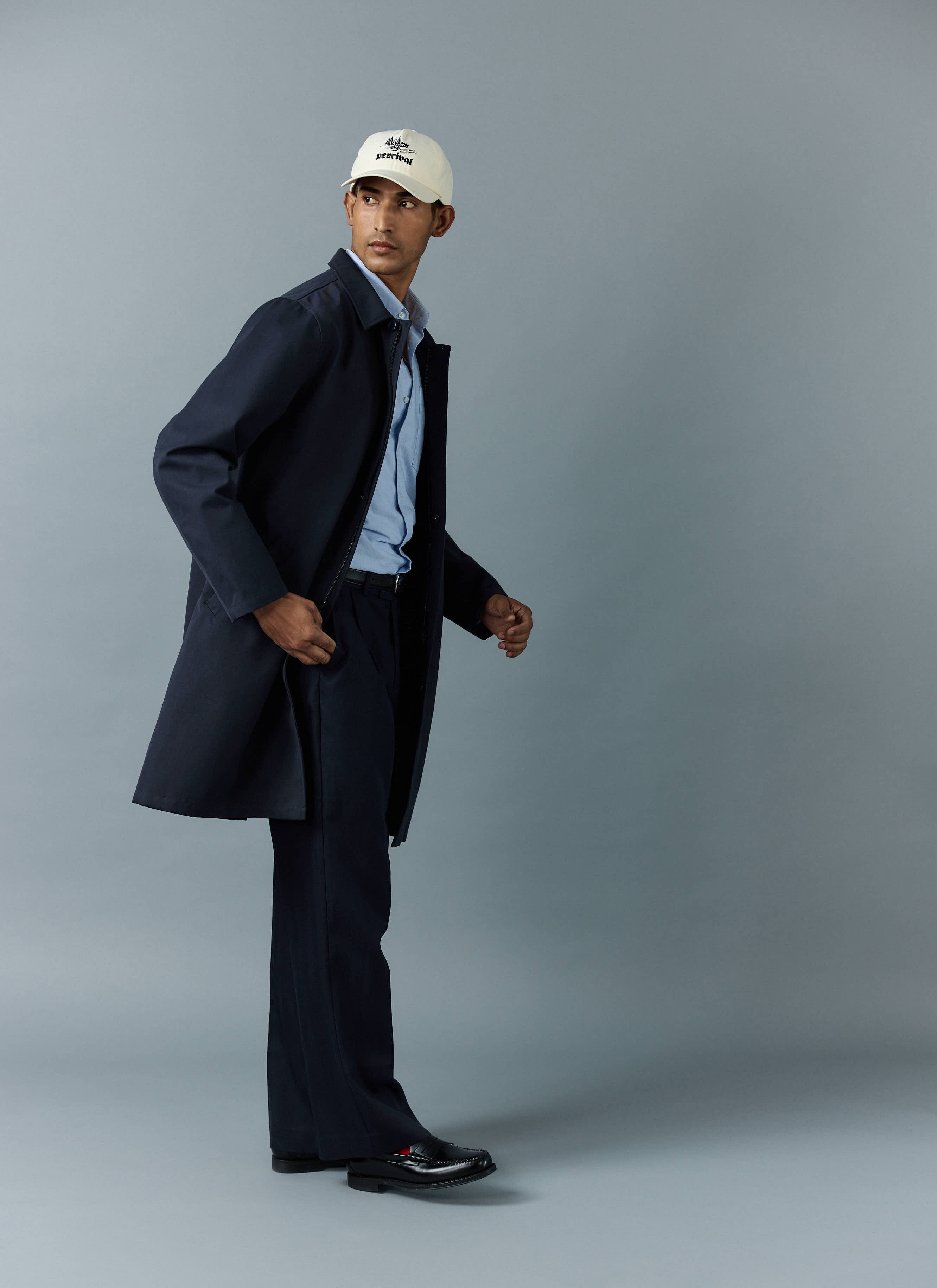 A model wearing the navy waterproof Greyson Trench Coat from Percival, styled with a blue shirt, trousers, and a cap.